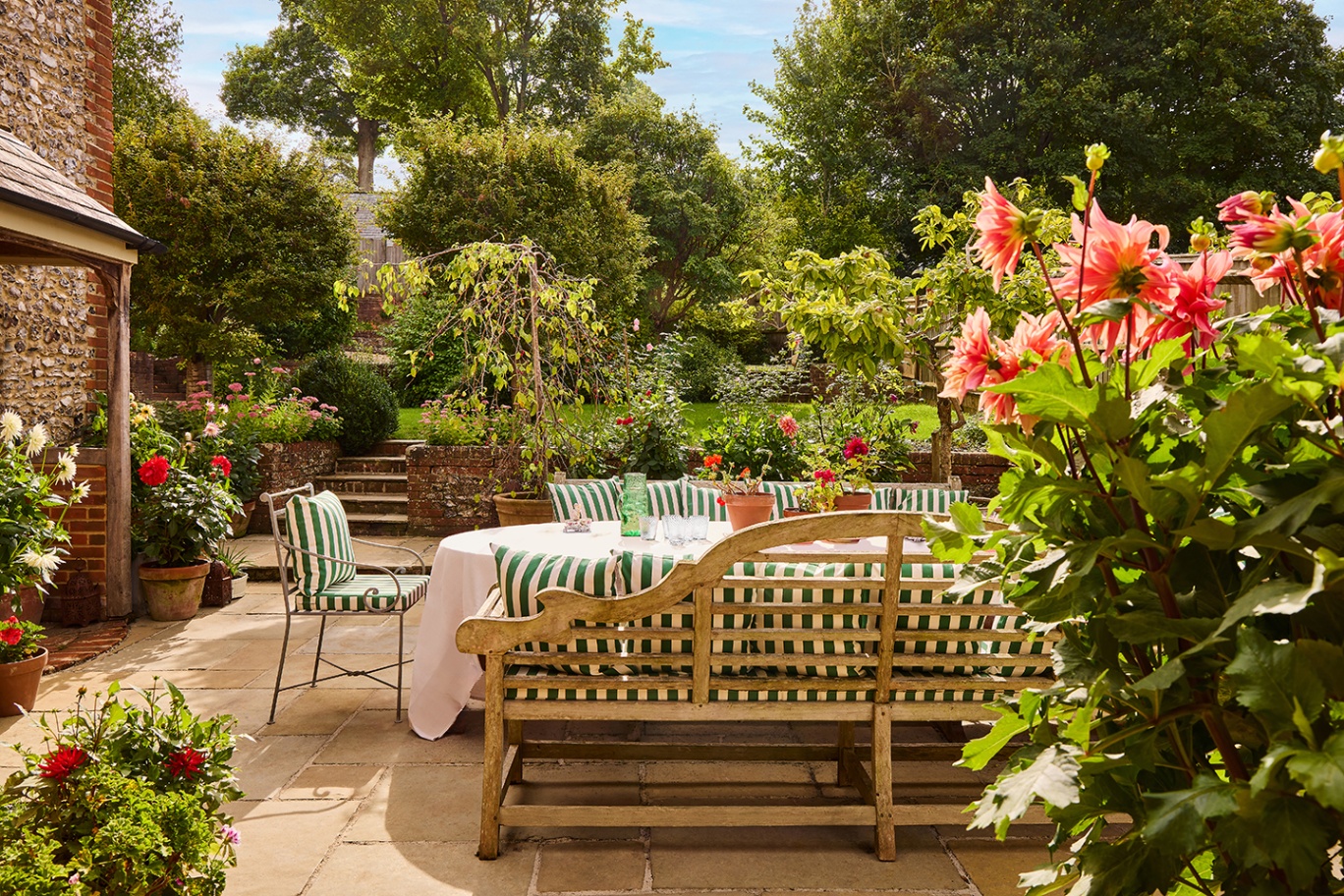 A large outdoor table dressed in a white tablecloth surrounded by metal chairs and wooden benches topped with striped cushions sits on a patio. In the background a lawn is visible and potted flowers are dotted around 