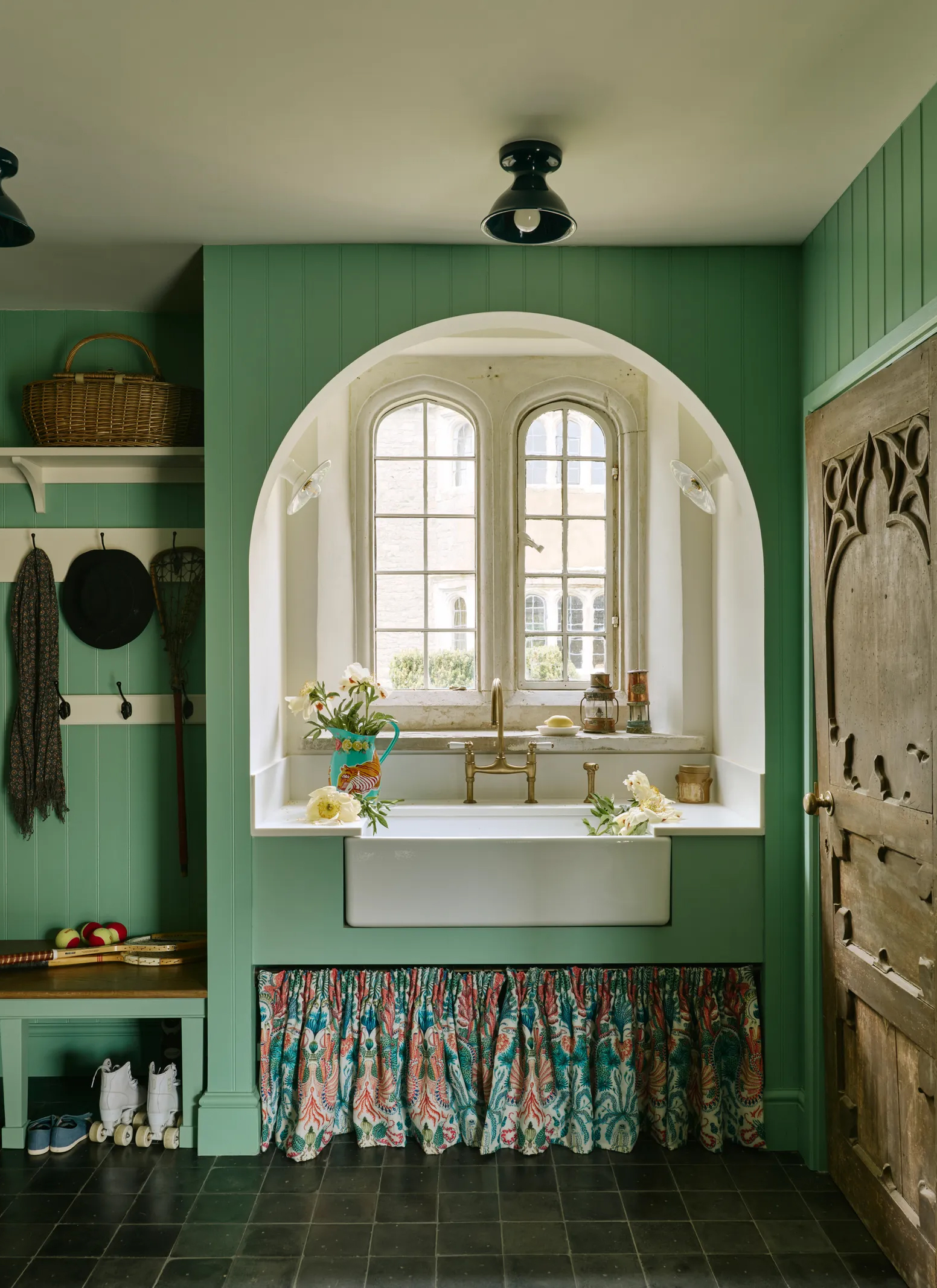 a boot room and utility room area with green painted walls and joinery, an arched window and a belfast sink with an orange and green floral sink skirt