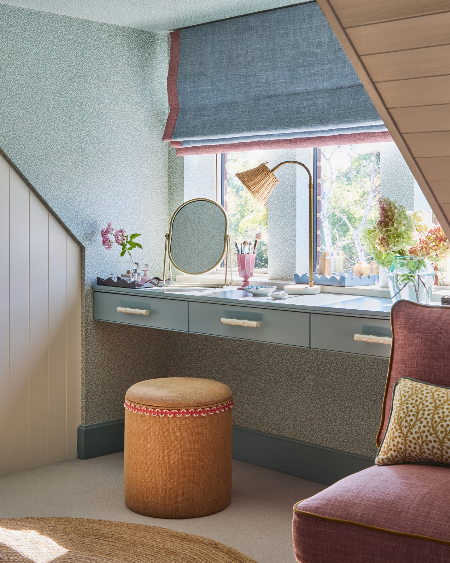 a bedroom corner with blue floral wallpaper, a blue floating dressing table with an oval mirror and a rattan lamp with an ochre upholstered stool