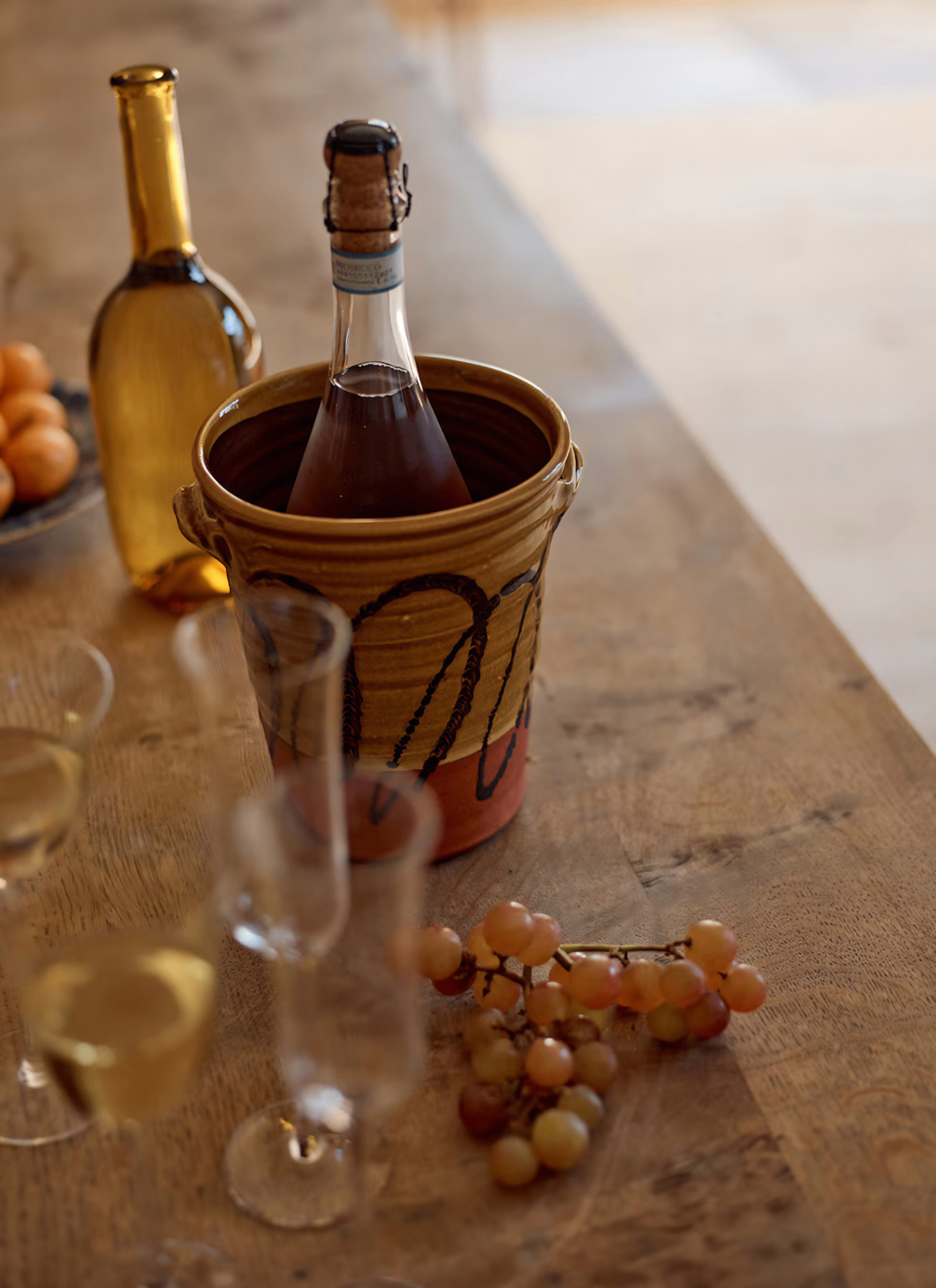 a close up of a wooden table with a patterned terracotta wine cooler with a bottle of wine inside, a bunch of red grapes and a collection of clear glassware
