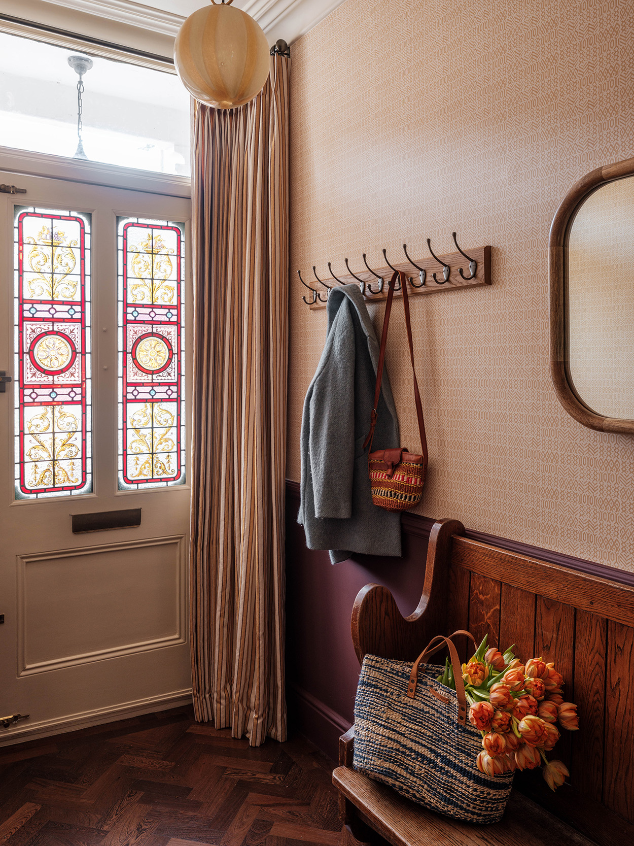 Hallway with traditional Victorian stained glass door with a pulled-back striped door curtain, the walls are covered in a rich aubergine paint on the lower half and a graphic wallpaper on the upper half, a striped glass globe pendant hangs above