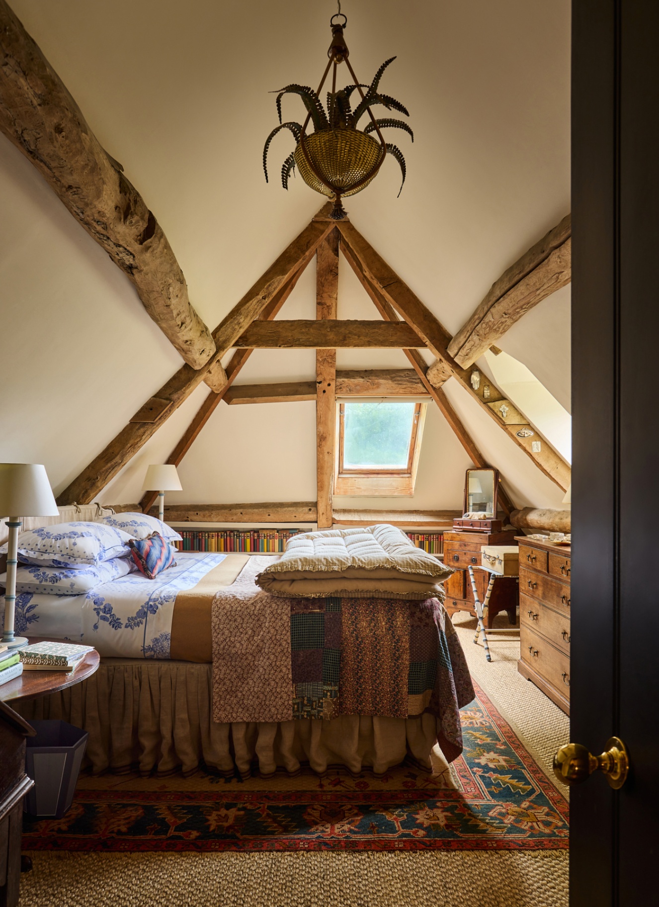 an eaves bedroom with white walls and wooden beams, a large ornate pendant light, antique wooden furniture and a large bed with floral bedding