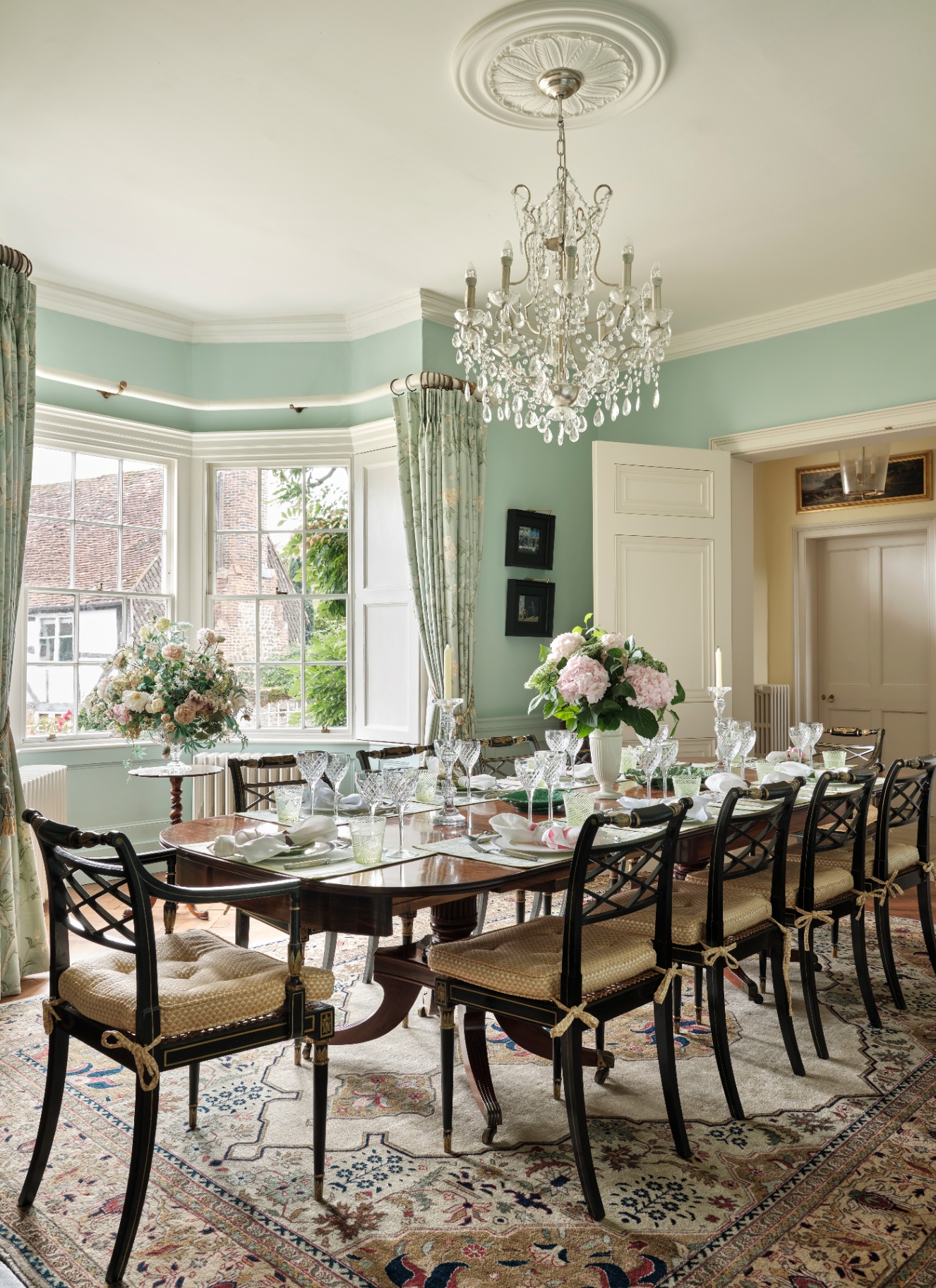 Light duck egg blue dining room in a historic period property with a large bay window, elegant crystal chandelier, long mahogany dining table, and classical black dining chairs