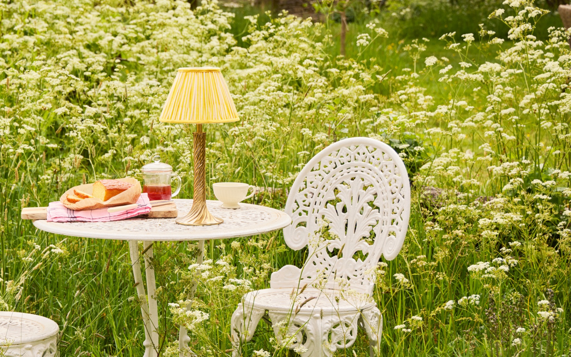 A GOLD AND YELLOW RECHARGEABLE LAMP ON A WHITE IRON BISTRO TABLE WITH CHAIRS, SURROUNDED BY LONG GRASS AND FLOWERS