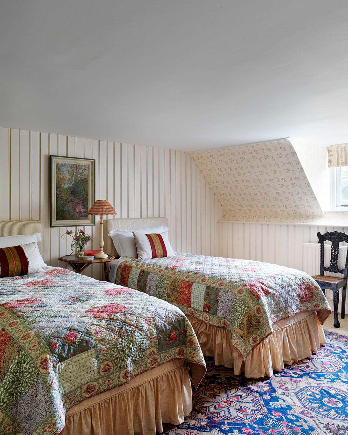 a cottage bedroom with white striped wallpaper, two twin beds with a patchwork patterned bedspread and pale pink bed valance sheets