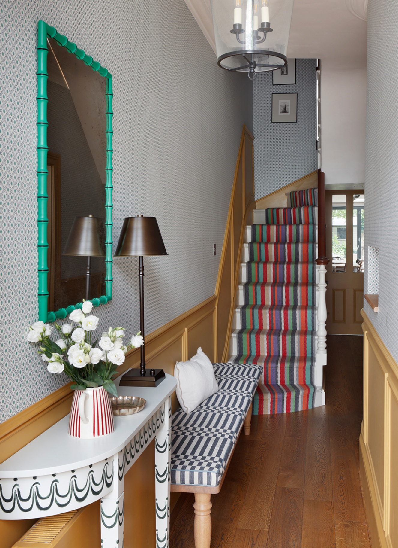 a hallway with a white console, a black lamp, a teal green mirror, stairs with a multicoloured stripe runner and yellow panelled walls