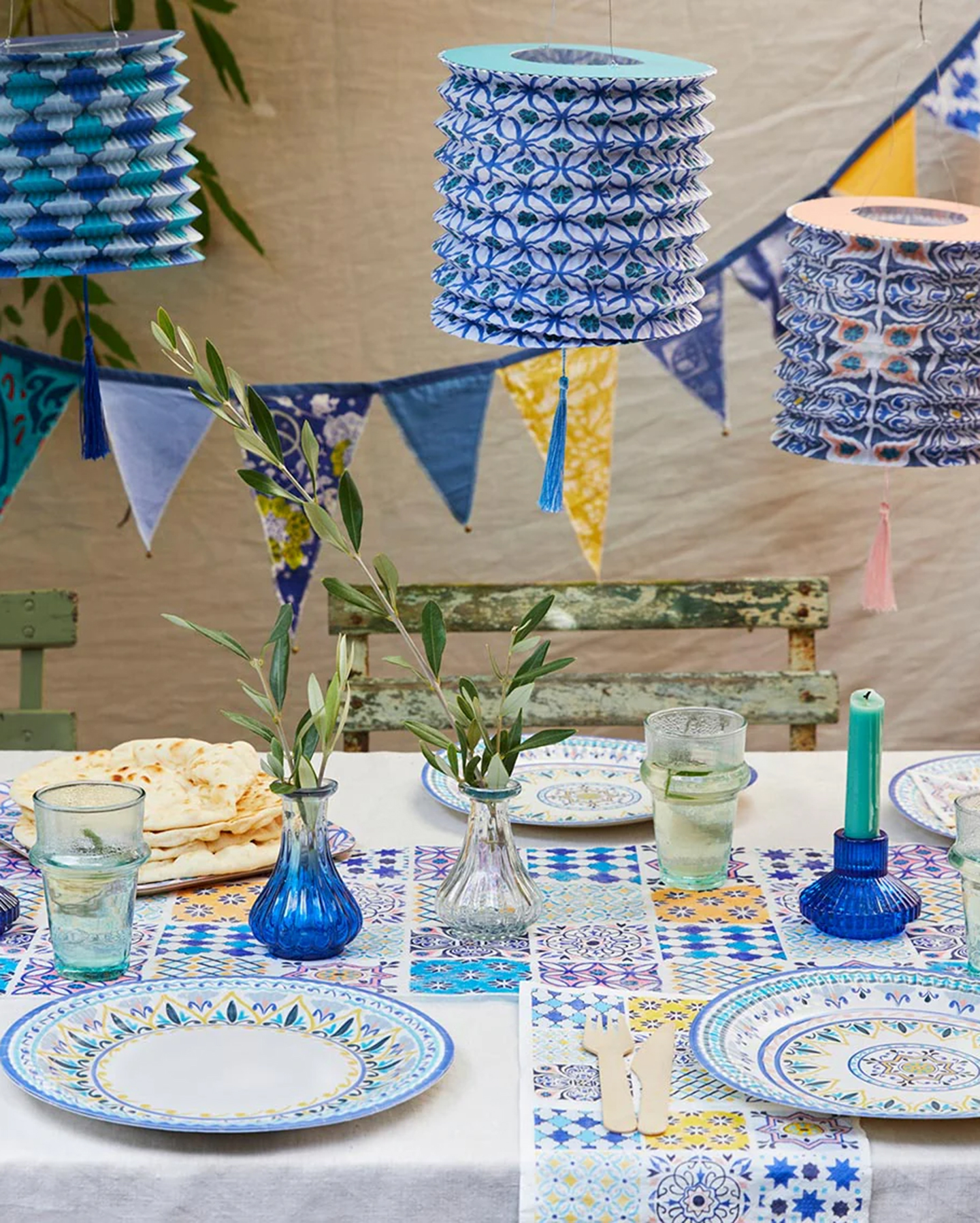 a dining table with blue and yellow tile pattened plates and table runners and blue patterned paper lanterns hanging above