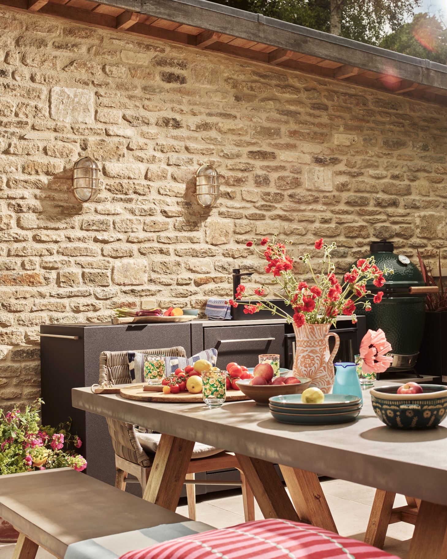an outdoor kitchen with a wooden table with a bench topped with plates and a terracotta jug with red flowers and an outdoor kitchen unit behind the table in front of a cotswold stone wall