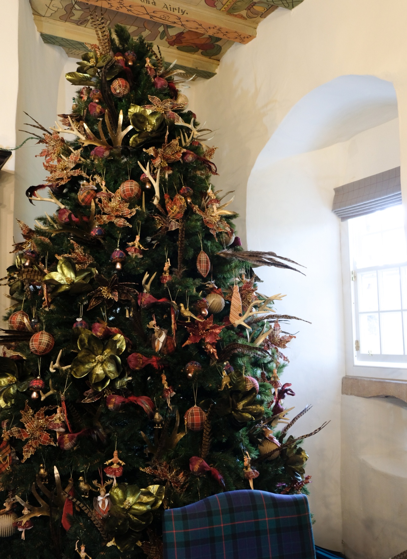 A grand, densely decorated Christmas tree filled with woodland ornaments, faux antlers, and tartan baubles next to an antique bookcase