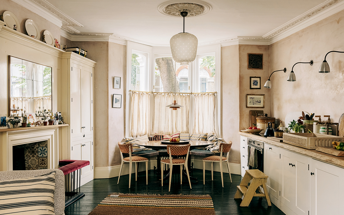 a kitchen with plaster walls and a run of lower level white cabinetry on the right, a large bay window in the centre where an oval table, three dining chairs and a built-in bench sit, and to the left if a fireplace and built-in tall cabinet