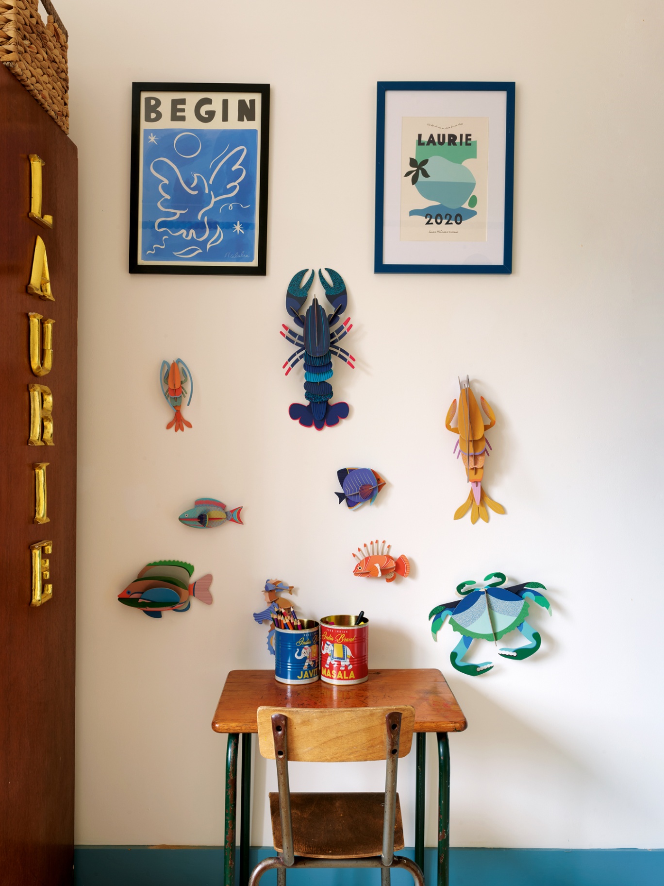 A close up of a childrens wooden desk and chair with two twins of pencils and pens on the desk. Above on a white painted wall is a collection of crafted cardboard sea creatures and personalised, colourful artworks