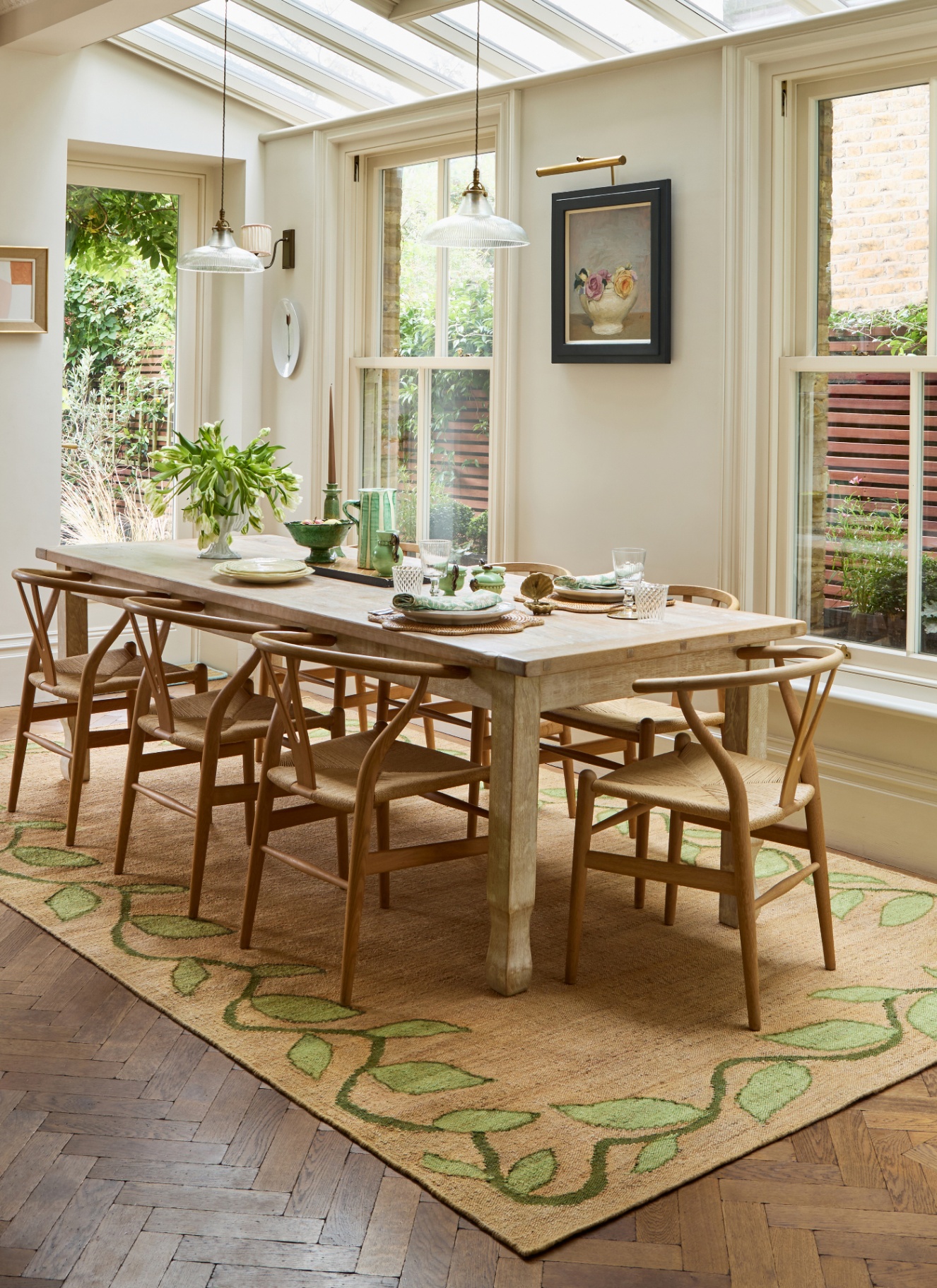 a dining room with white walls, two glass pendant lights over a long wooden table and chairs and a jute rug with green details
