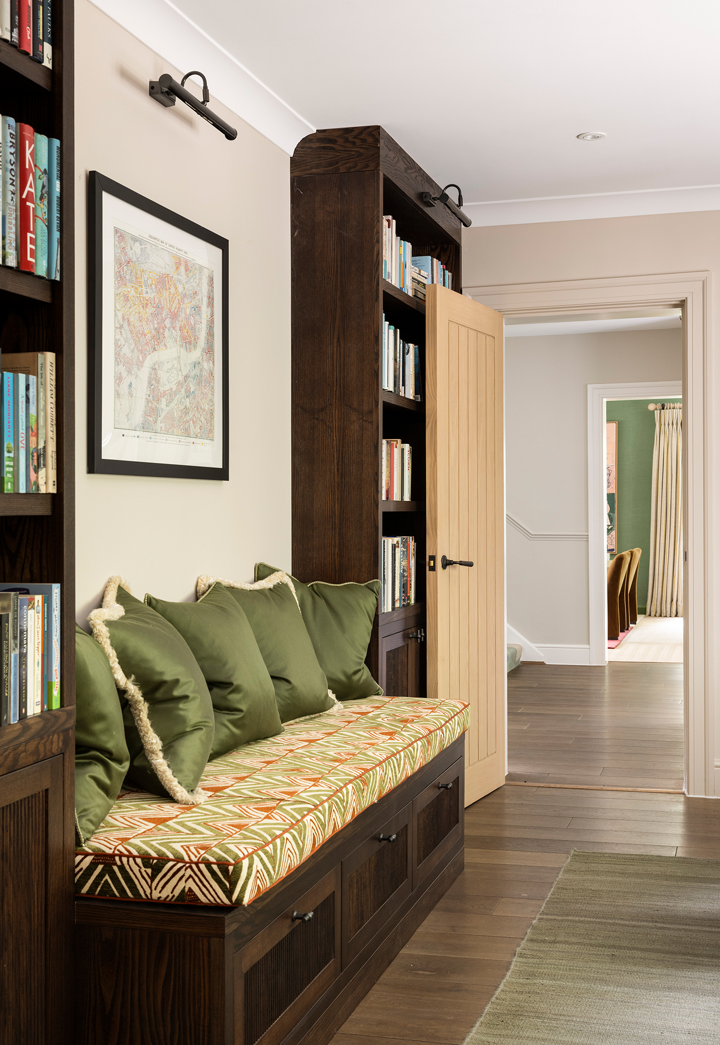 a close up of a built in bench with a graphic upholstered seat cushion topped with green fringed cushions. Above hangs a framed map artwork and picture light, and either side are built-in wooden bookcases filled with books