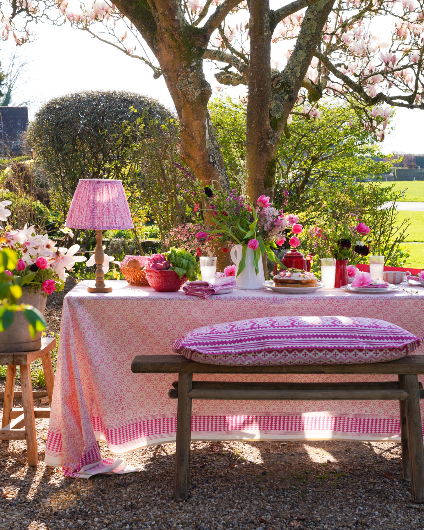an outdoor spring tablescape with pink tablecloth, pink flowers, a pink cushion on the bench and a pink pleated outdoor lamp