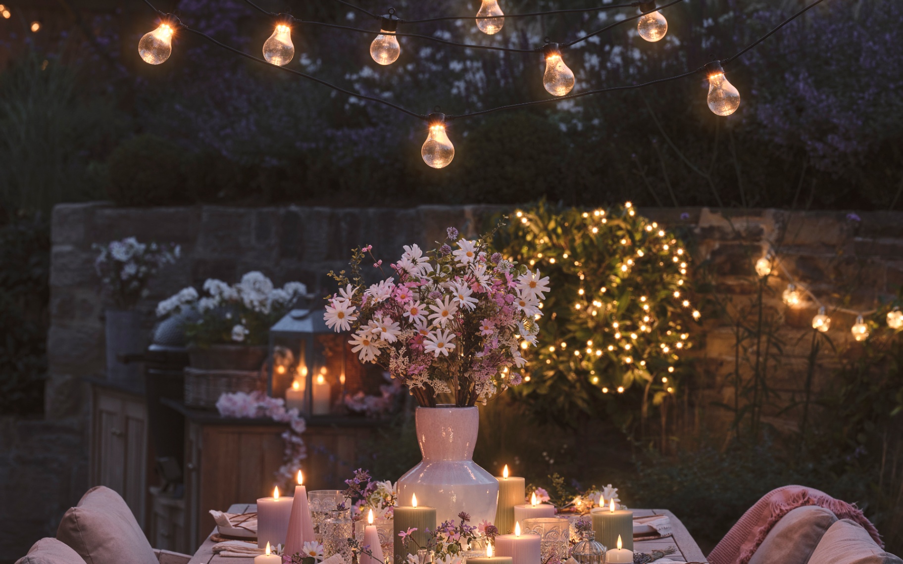 an outdoor dining table at light with led candles and a vase of flowers with festoon lights hanging above it