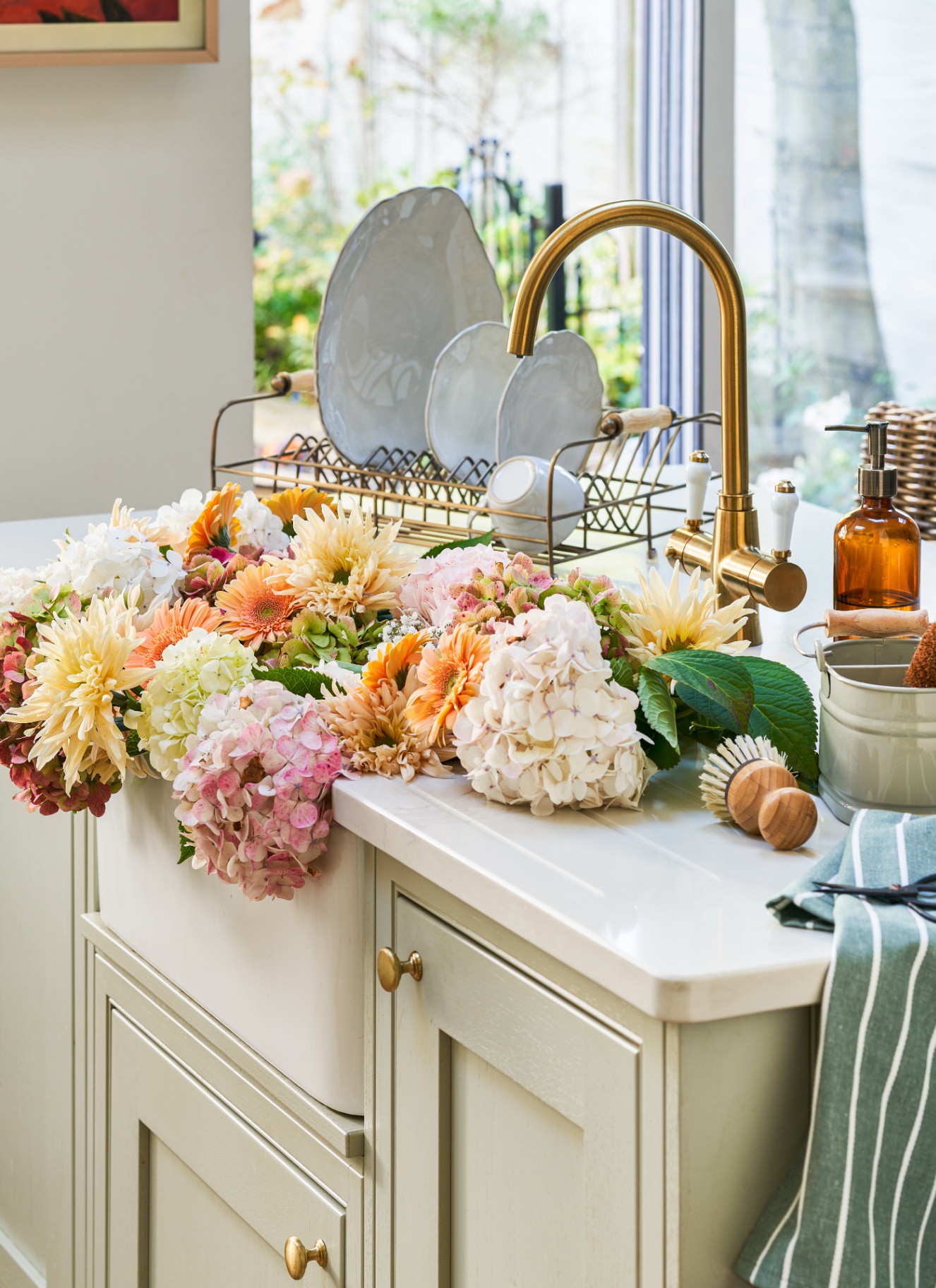 a kitchen sink area with a bunch of flowers in the sink, sage green cabinets and a wire plate drying rack