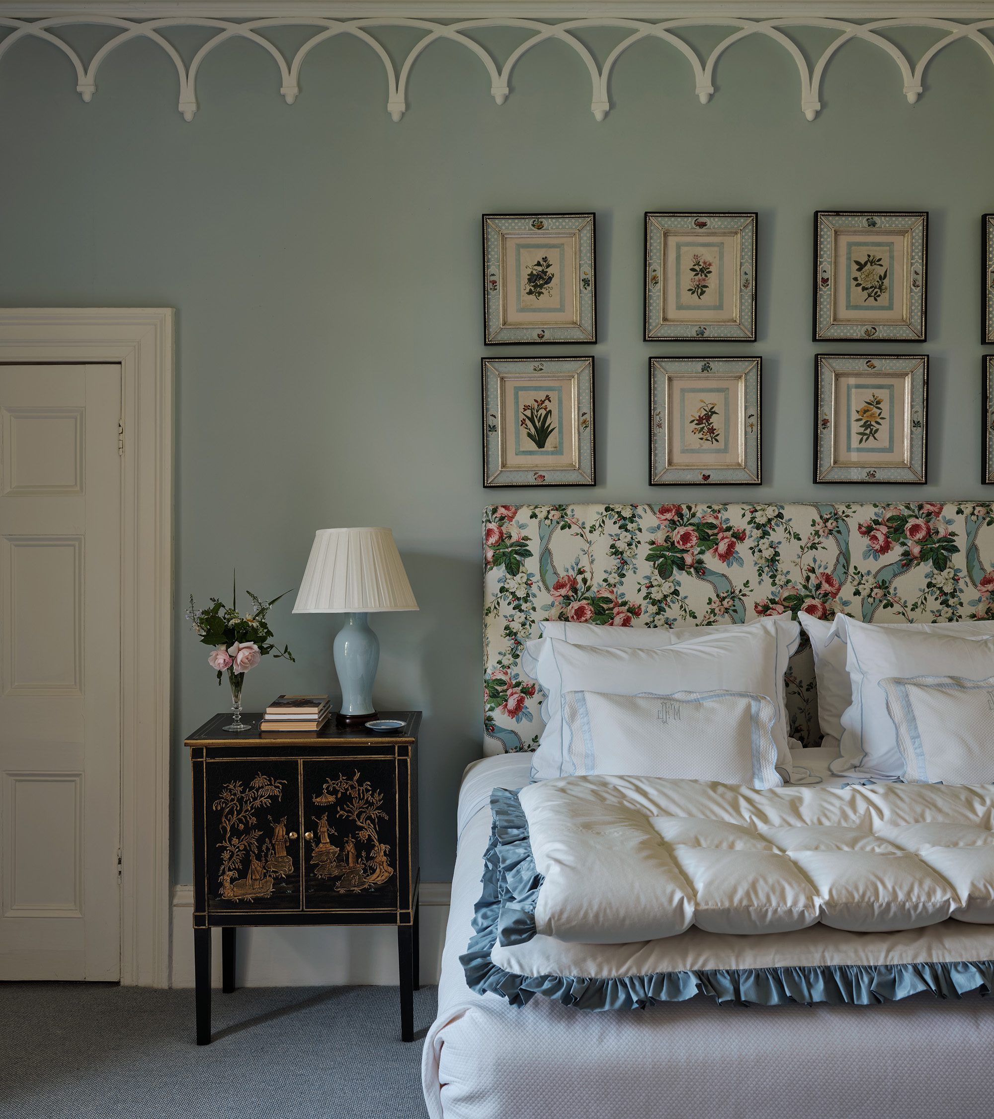 a traditional bedroom with pale blue walls, an antique bedside table, a set of 6 art prints above the bed, and a bed with white bedding and a floral headboard