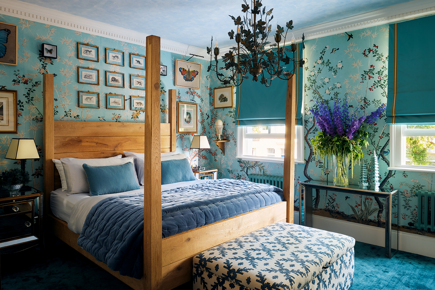 a bedroom with a large wooden four poster bed in the middle, with an upholstered ottoman at the end. The walls are covered in a botanical mural wallpaper and a metal botanical chandelier hangs in the middle of the room. Blue carpet lines the floor