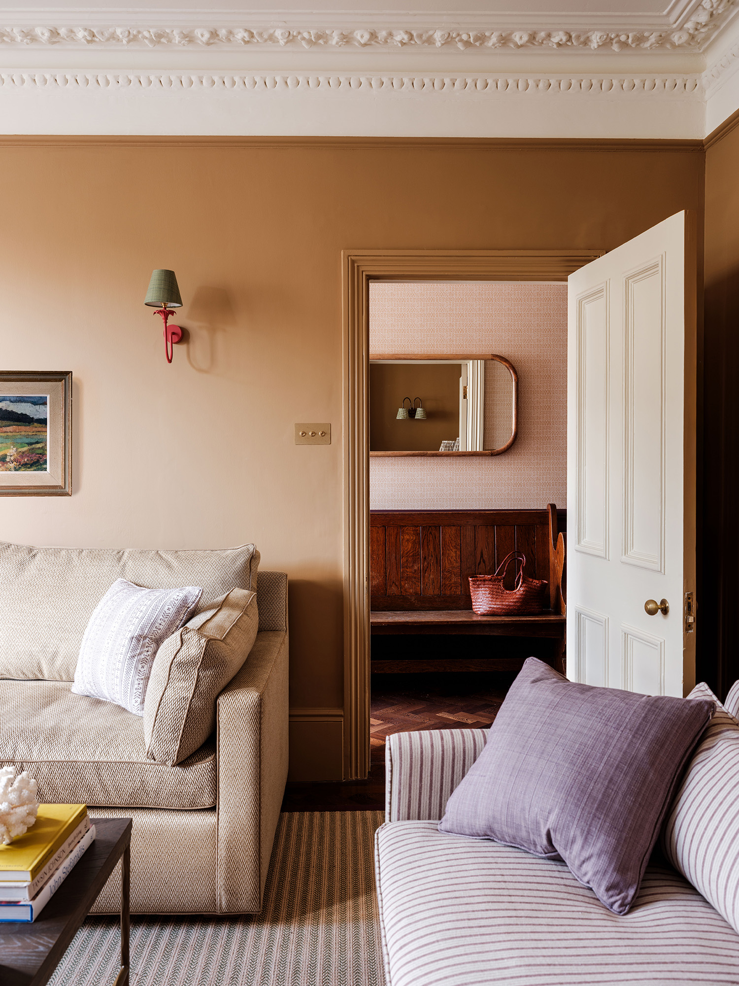 Victorian living room with ornate cornicing and caramel brown painted walls, two sofas are in the foreground, each upholstered in small-print fabrics, and in the background an open door looks through to the hall with a large wooden bench and curved mirror