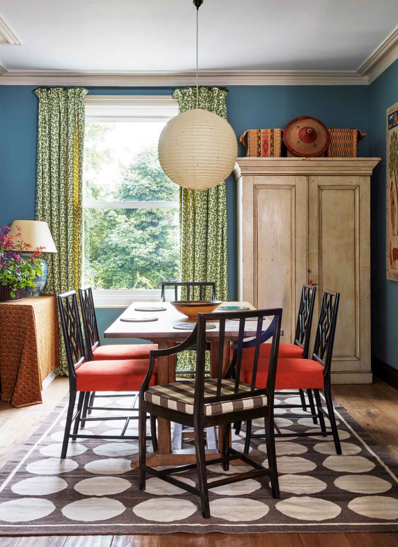 Bright blue dining room featuring dark wood dining table, orange upholstered chairs, patterned geometric rug, botanical curtains, and a large paper lantern light fixture.