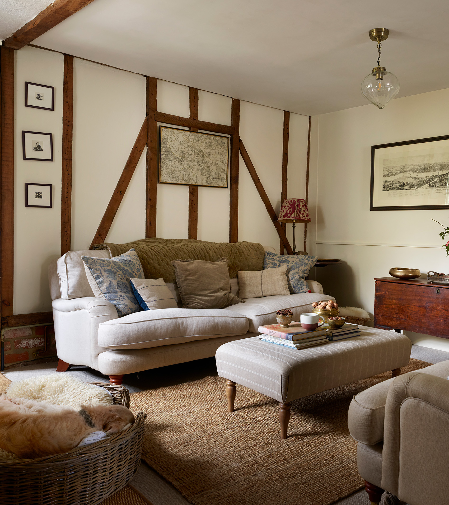 a white living room with wooden beams, a neutral sofa suite with a matching ottoman in the middle and a wicker dog basket on the bottom left with a sleeping golden retriever inside