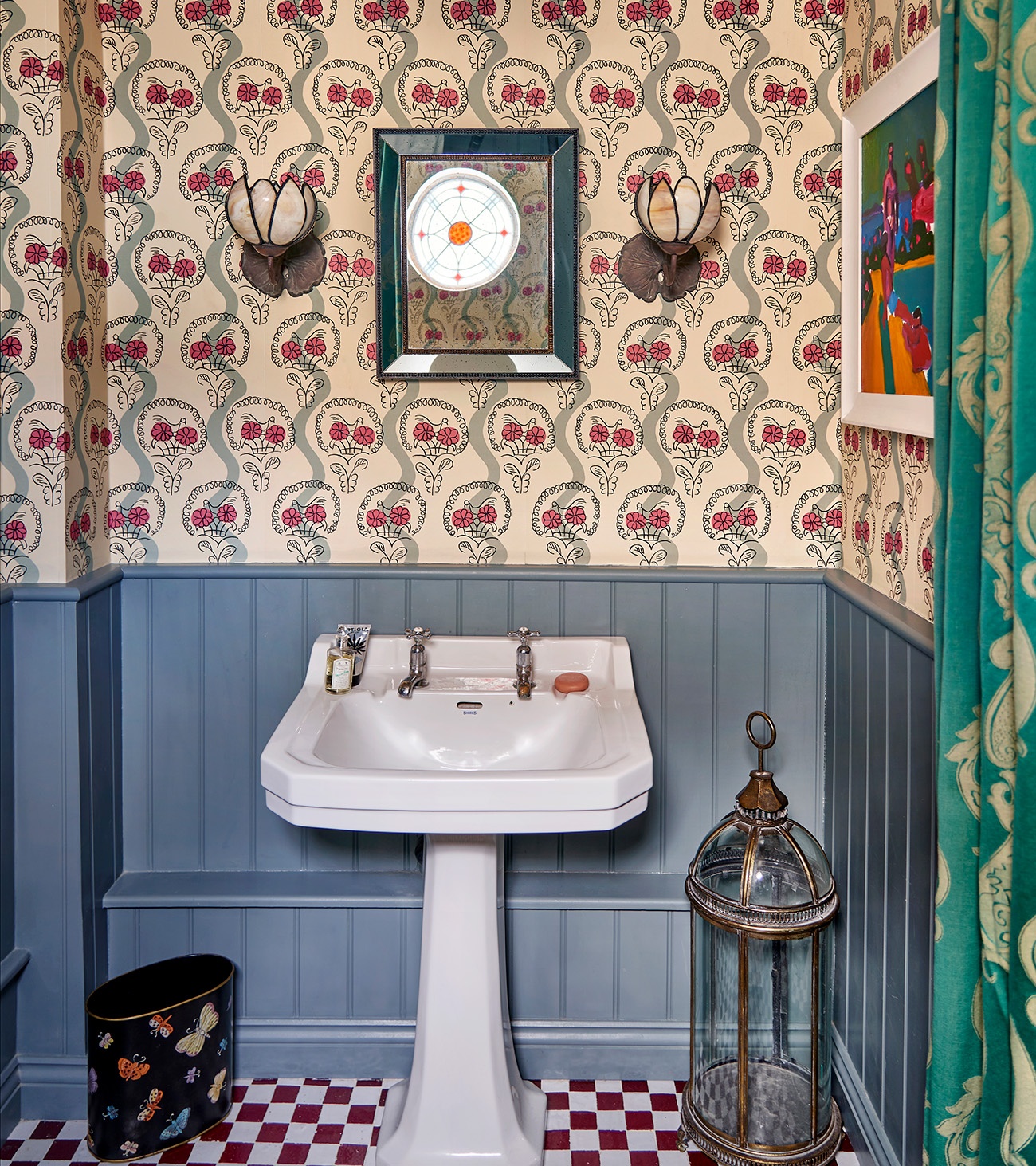 a powder room with cornflower blue paneling on the bottom half of the wall and cream and pink botanical wallpaper on top, a small rectangular bathroom mirror with two glass sconces either side, a white sink and a large lantern next to it