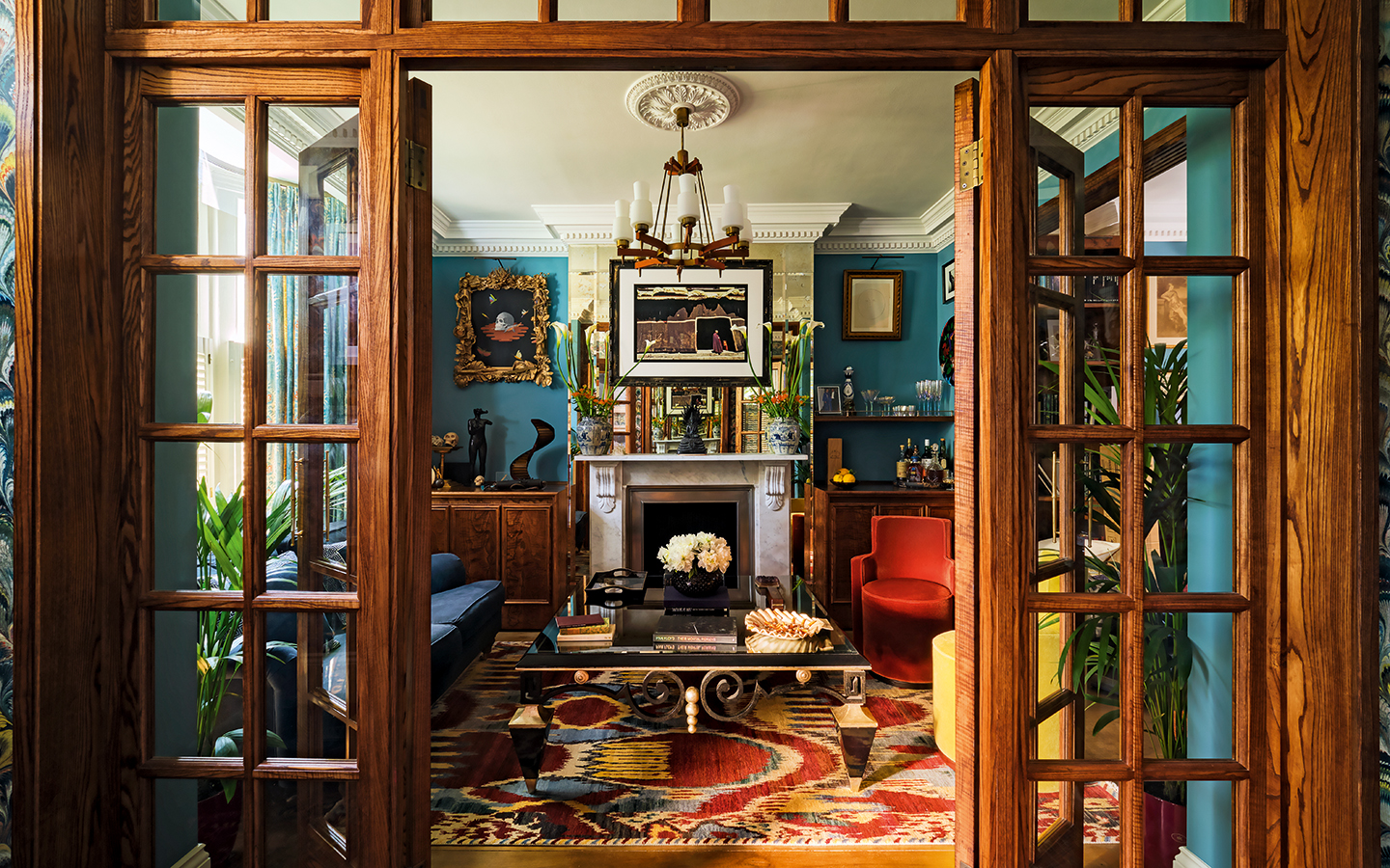 A view through glass-paned wooden internal doors to a rich blue living room with colourful sofa and accent chairs, a large ornate coffee table and a vibrant rug underneath. A fireplace is on the back wall and lots of art is on display