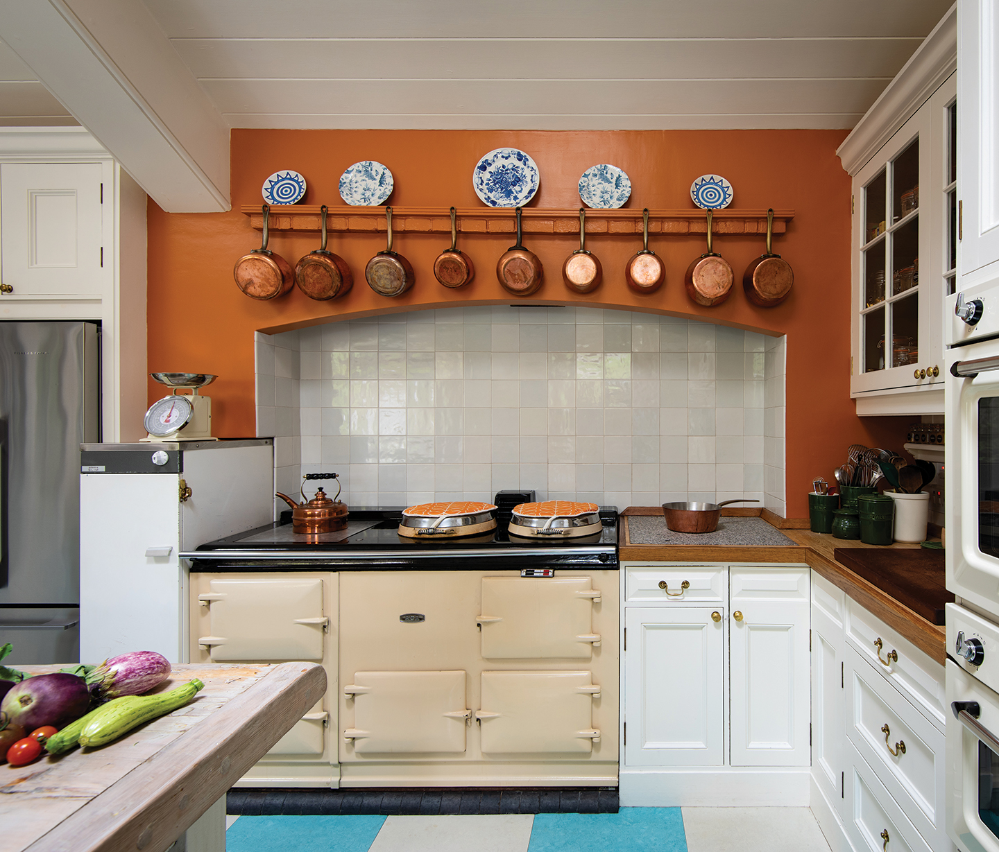 a kitchen with burnt orange walls and a blue and white checkerboard floor. The cabinets are white with wooden worktops and there is a cream coloured aga with a copper kettle atop it and white tiles behind.