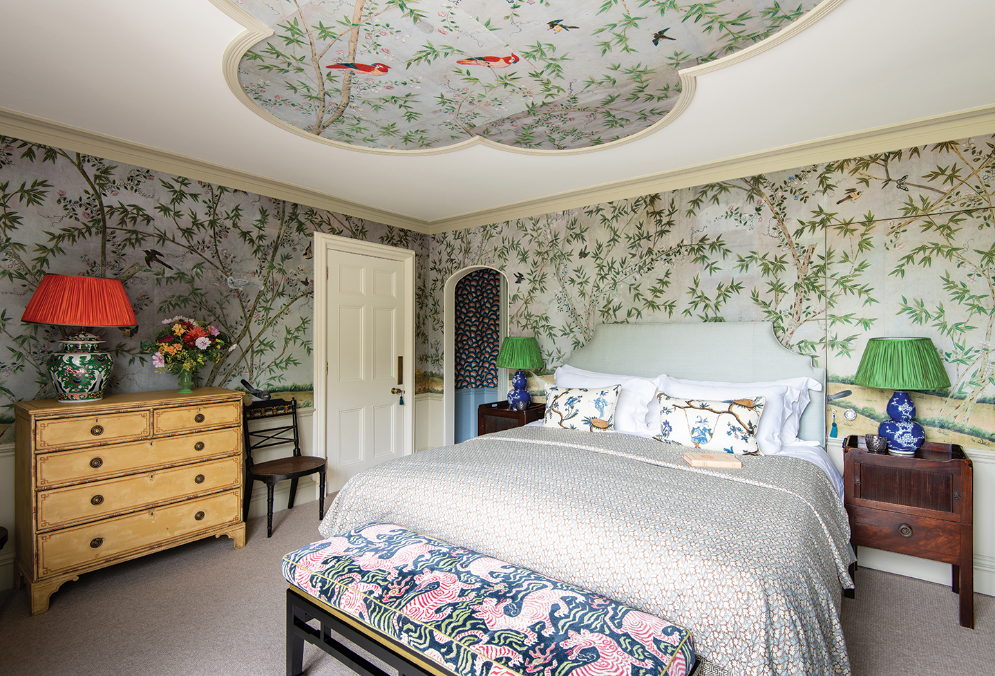 a large bedroom with walls and a quatrefoil design on the ceiling covered in chinoiserie-style wallpaper. A large double bed is flanked by wooden bedside tables with green and blue table lamps and at the end is a bench upholstered in a tiger print fabric