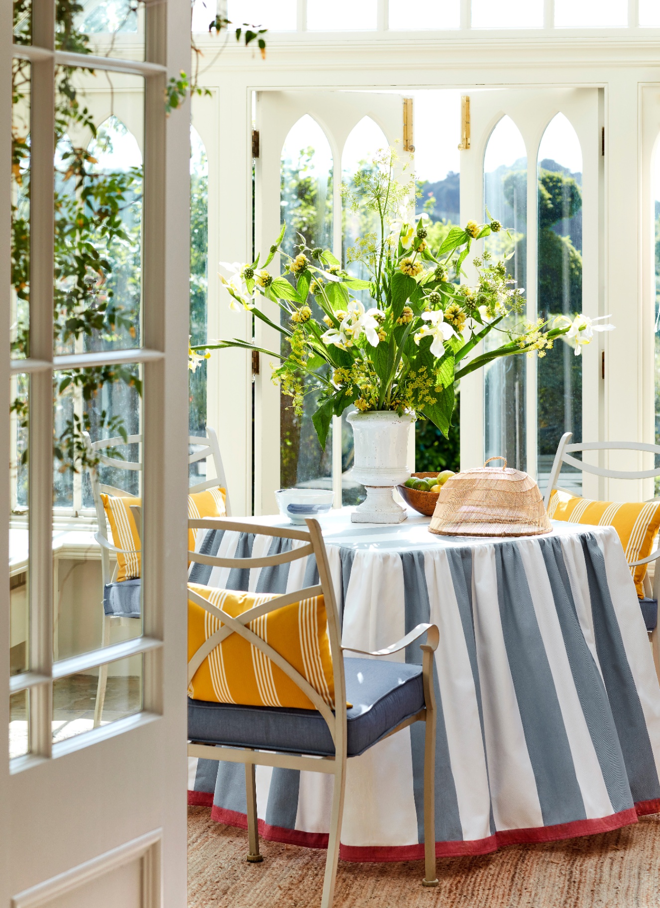 a conservatory with a round table covered in a blue and white striped table cloth and a large white vase with tall yellow and green flowers