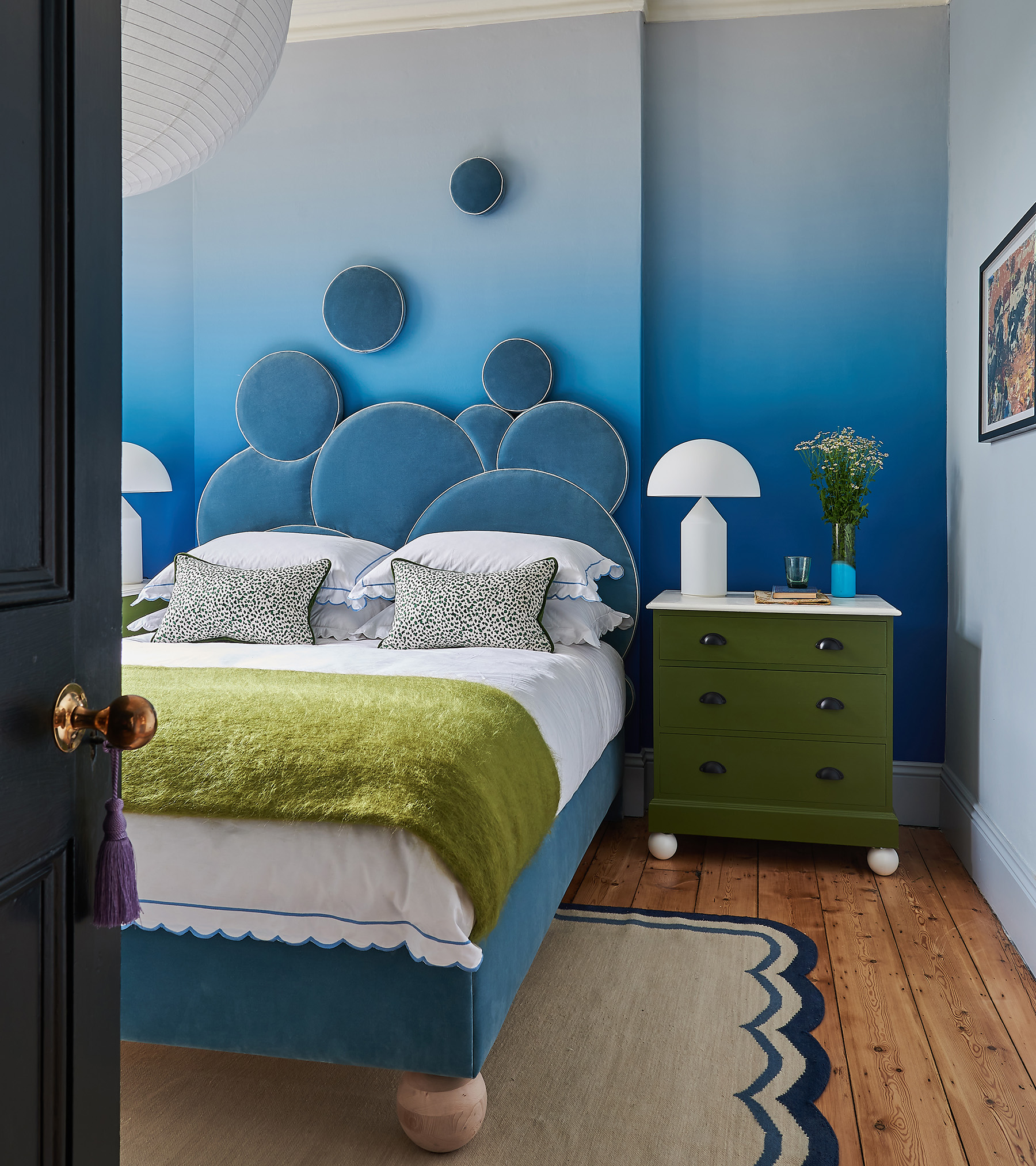 a bold blue bedroom with a circle arty headboard that trails up the wall, a blue and white ombre wall, a double bed with green throw, a green bedside table and a scallop rug