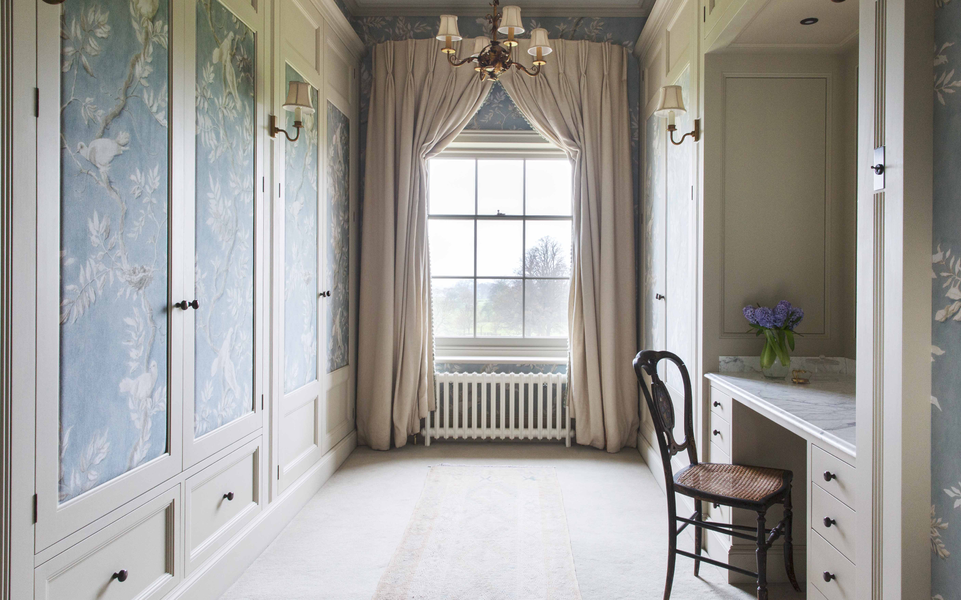 a dressing room with pale blue floral wardrobe doors, a large mirror at the back of the room and a built in dressing table to the right with a wooden chair