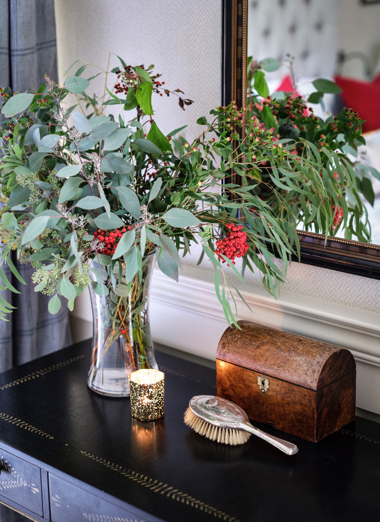 A glass vase filled with eucalyptus and clusters of bright red berries on a dark, patterned console table with a vanity brush, small chest, and a candle in front of a mirror.