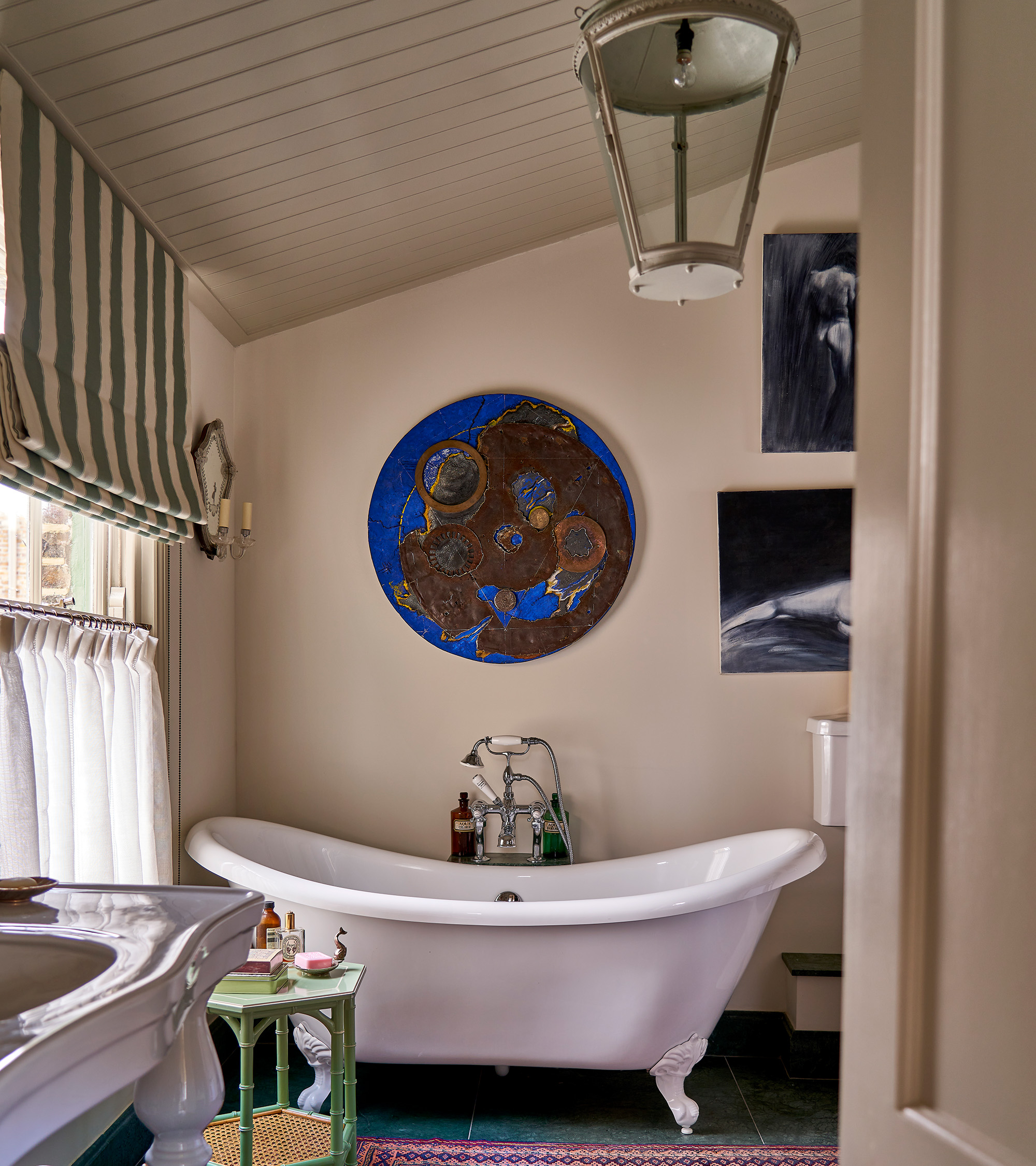 a main bathroom with a white roll top bath, a blue and brow circular wall art above it, a large window with a stiped blind and a large lantern style pendant on the ceiling