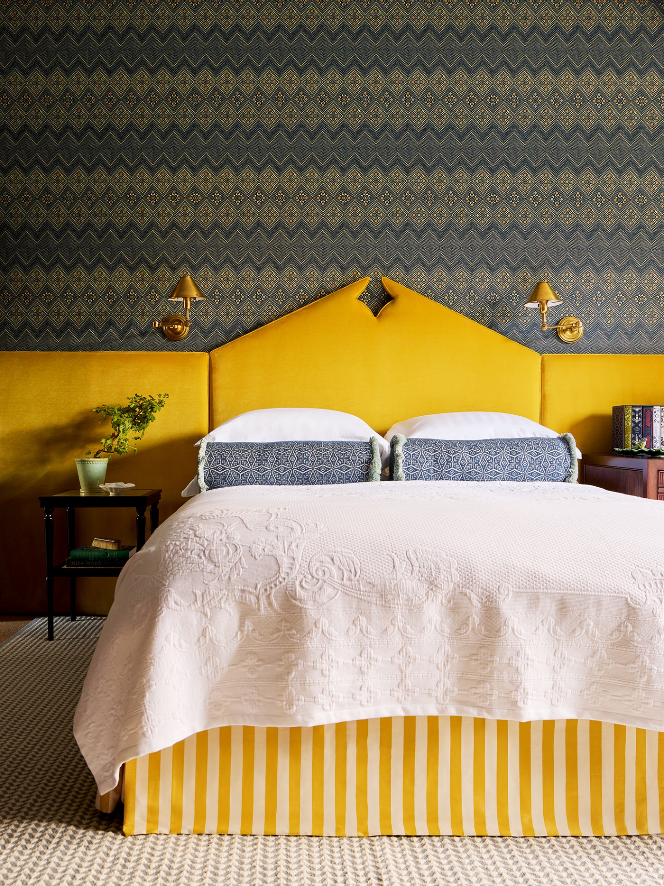 A bedroom with blue patterned wallpaper, a decorative yellow velvet headboard that extends along the back wall. The bed is dressed with white linens, blue bolster cushions and has a yellow and white stripe valance. Two brass wall lights are fixed above