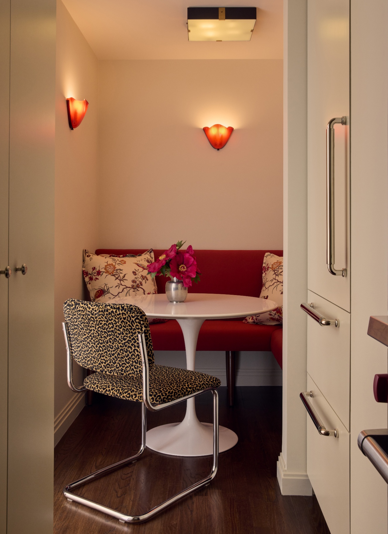Compact kitchen dining alcove with a dark red built-in banquette, white tulip pedestal table, leopard print chair, and warm wall sconces