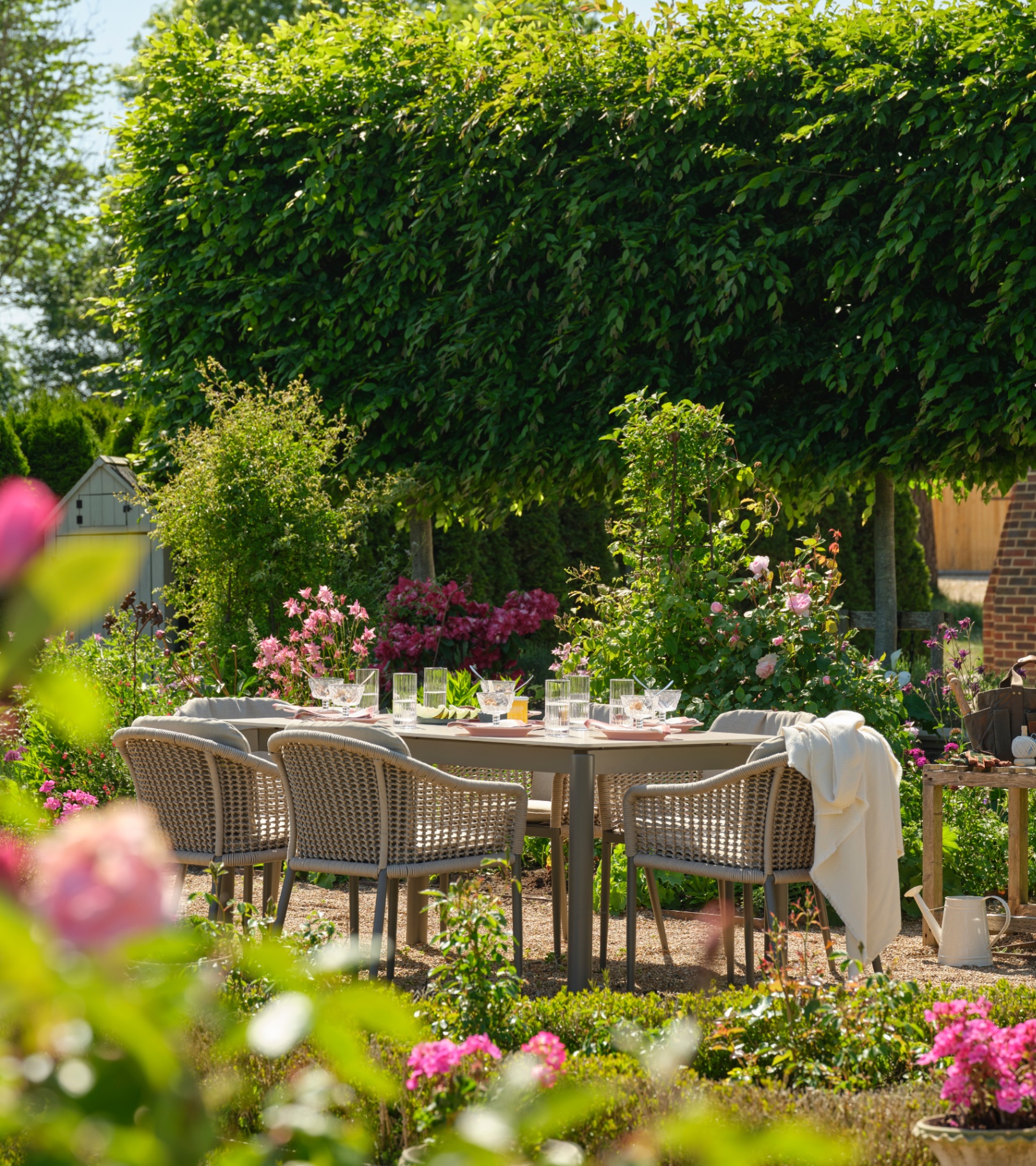 a set of grey rattan outdoor dining furniture on a patio surrounded by greenery and colourful flowers