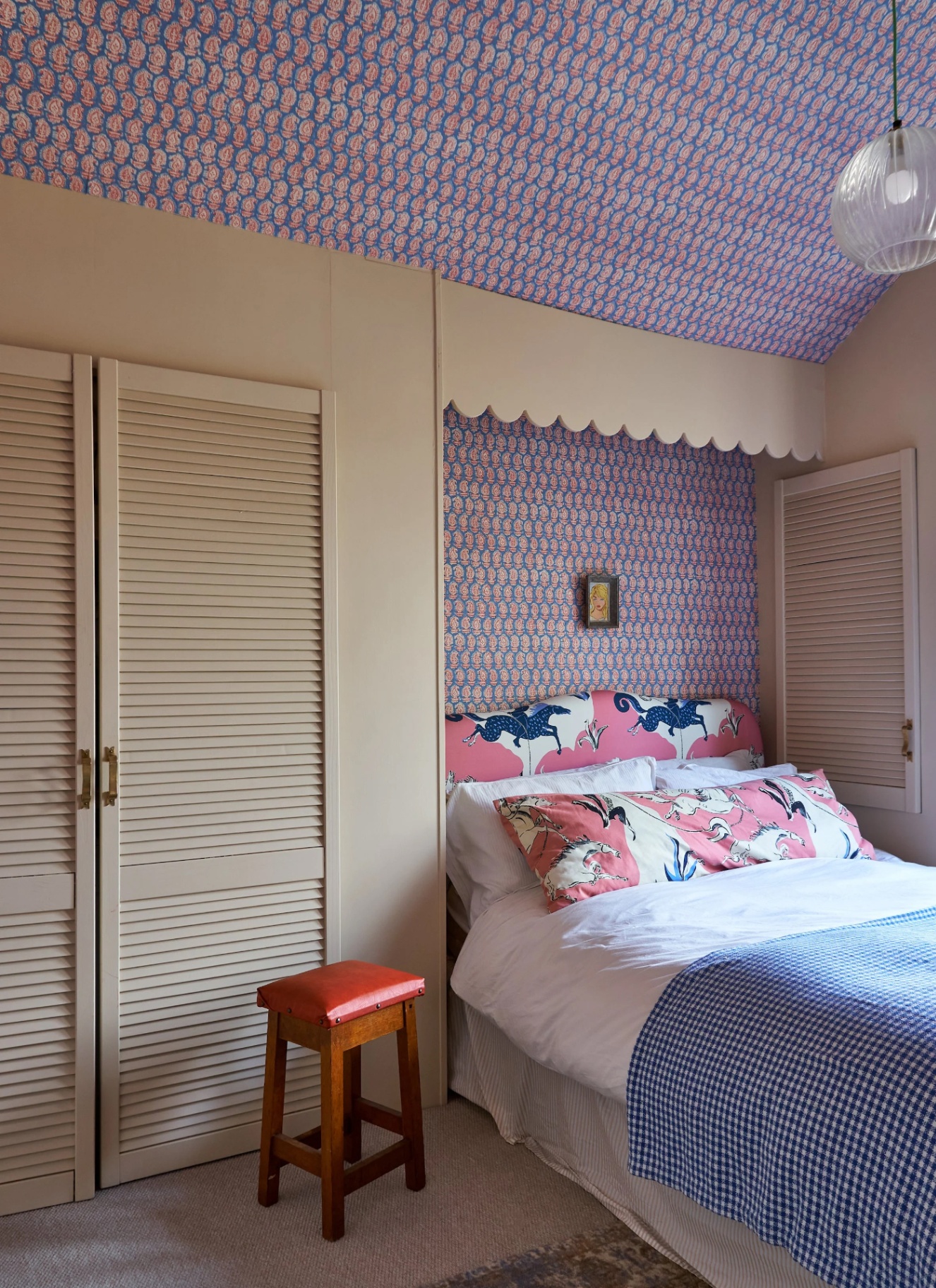 a bedroom with a purple and blue patterned wallpapered ceiling and wall behind the headboard, a bed with a pink upholstered headboard and blue gingham throw and beige cupboards