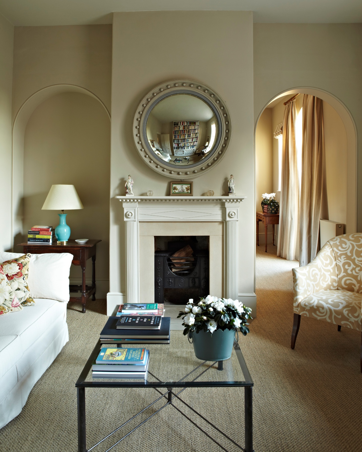 a neutral living room with a large circle mirror above a marble fireplace, a black and glass square coffee table with books and a pot of white roses and a white linen sofa