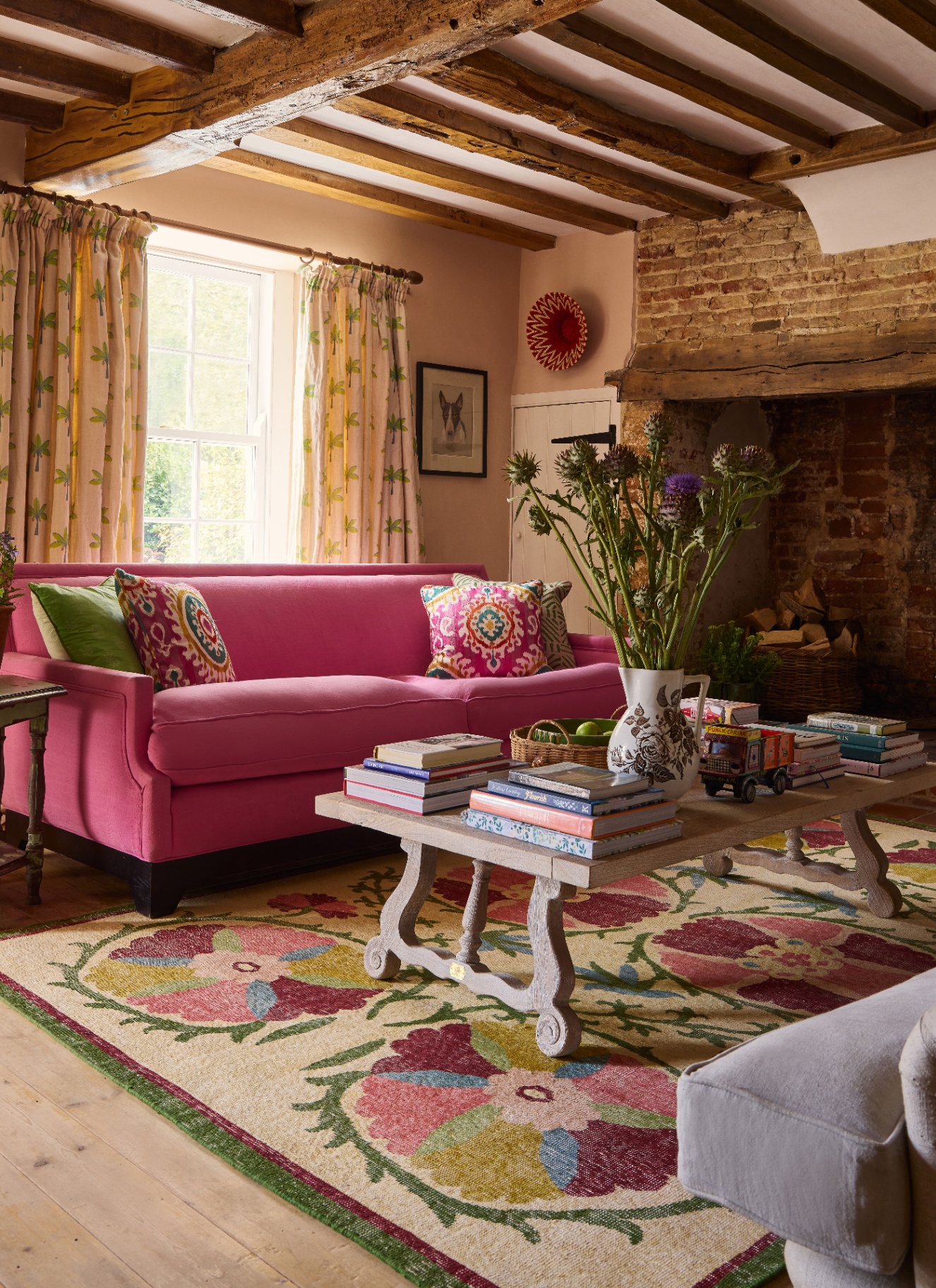 A rustic cottage sitting room featuring exposed ceiling beams, an exposed brick fireplace, a bright pink sofa, patterned floral curtains, and a colourful floral patterned rug under a wooden coffee table.