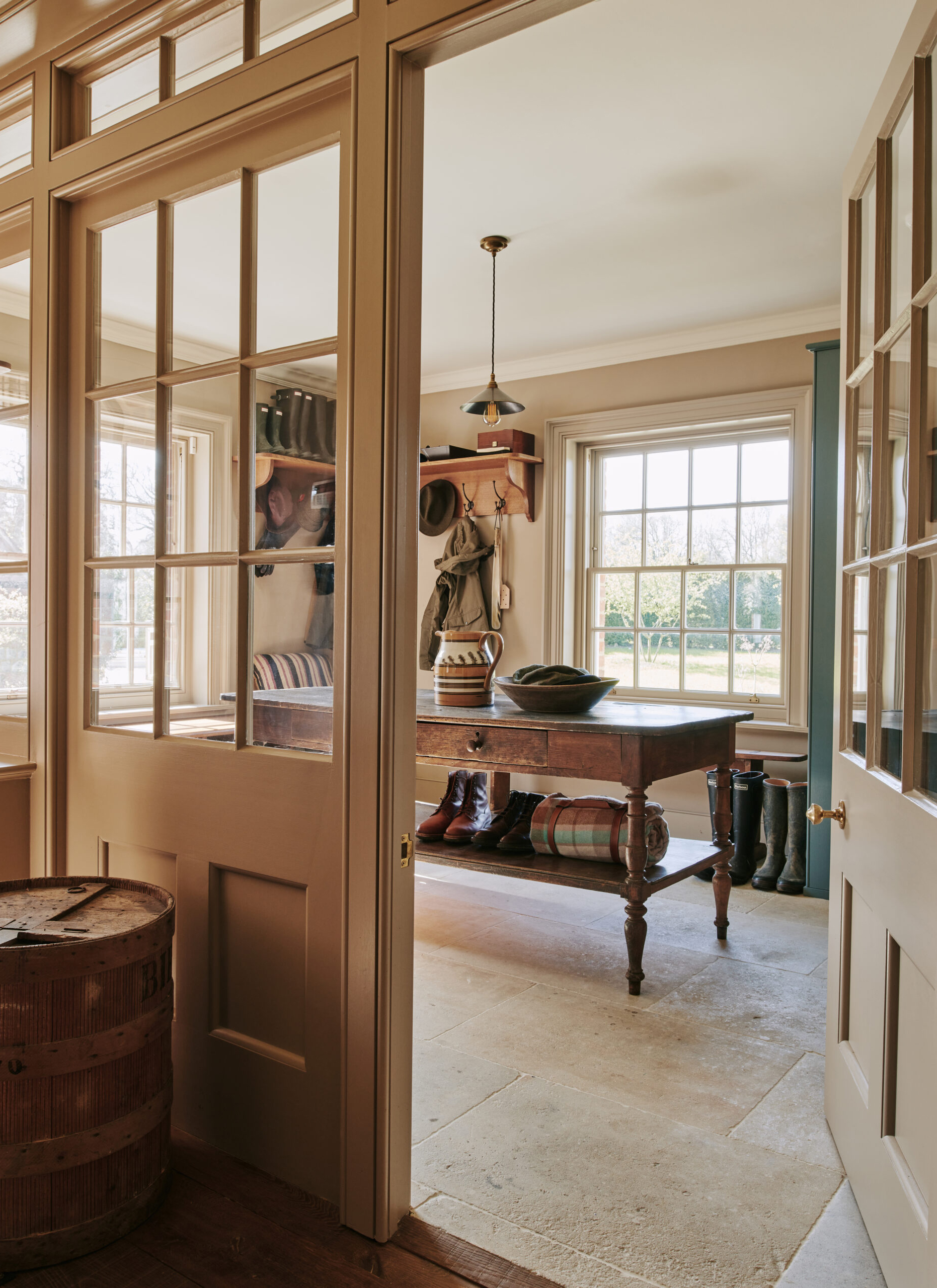 a boot room with cream stone floors, a large central console table with a blanket and a pair of boots under it and shelves with coats hanging and a row of wellington boots