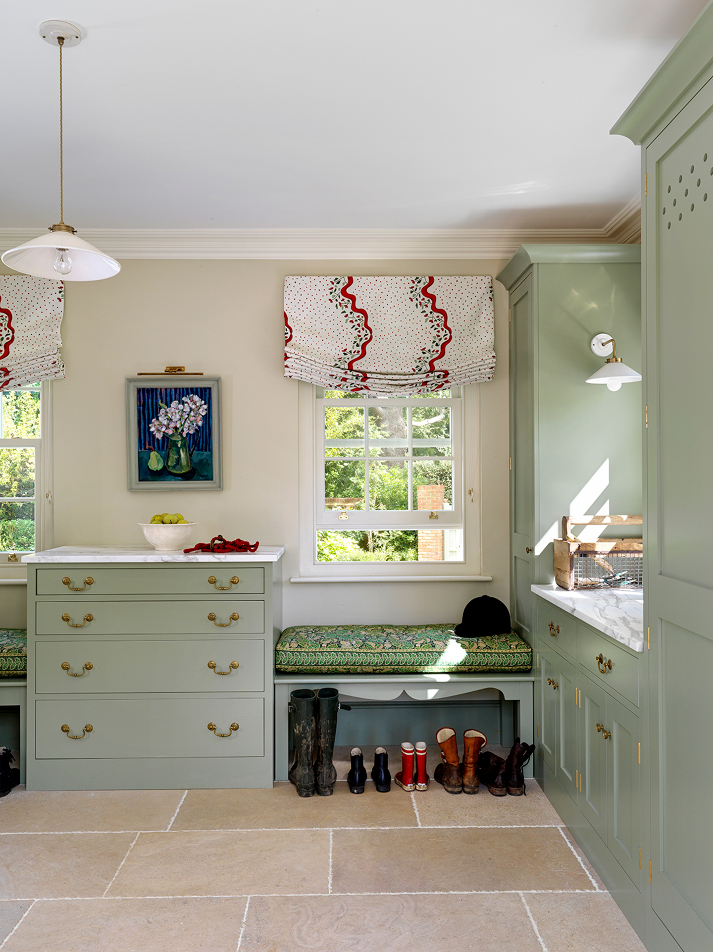 A boot room with flagstone flooring, soft green cabinetry, including tall cupboards and lower cabinets with drawers, and relaxed blinds dressing sash windows