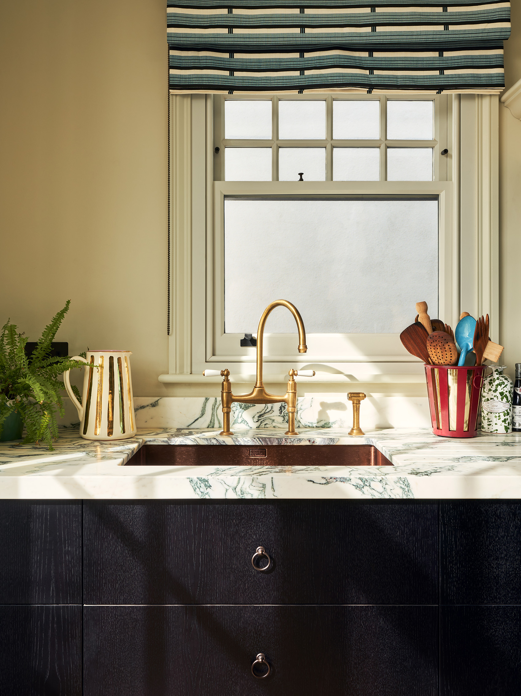 a close up of a copper kitchen sink set in a veined white marble worktop and black wood cabinet with a bronze ring pull, surrounded by a jug with gold details and a utensil holder, and above is a window with a blue and white fabric blind
