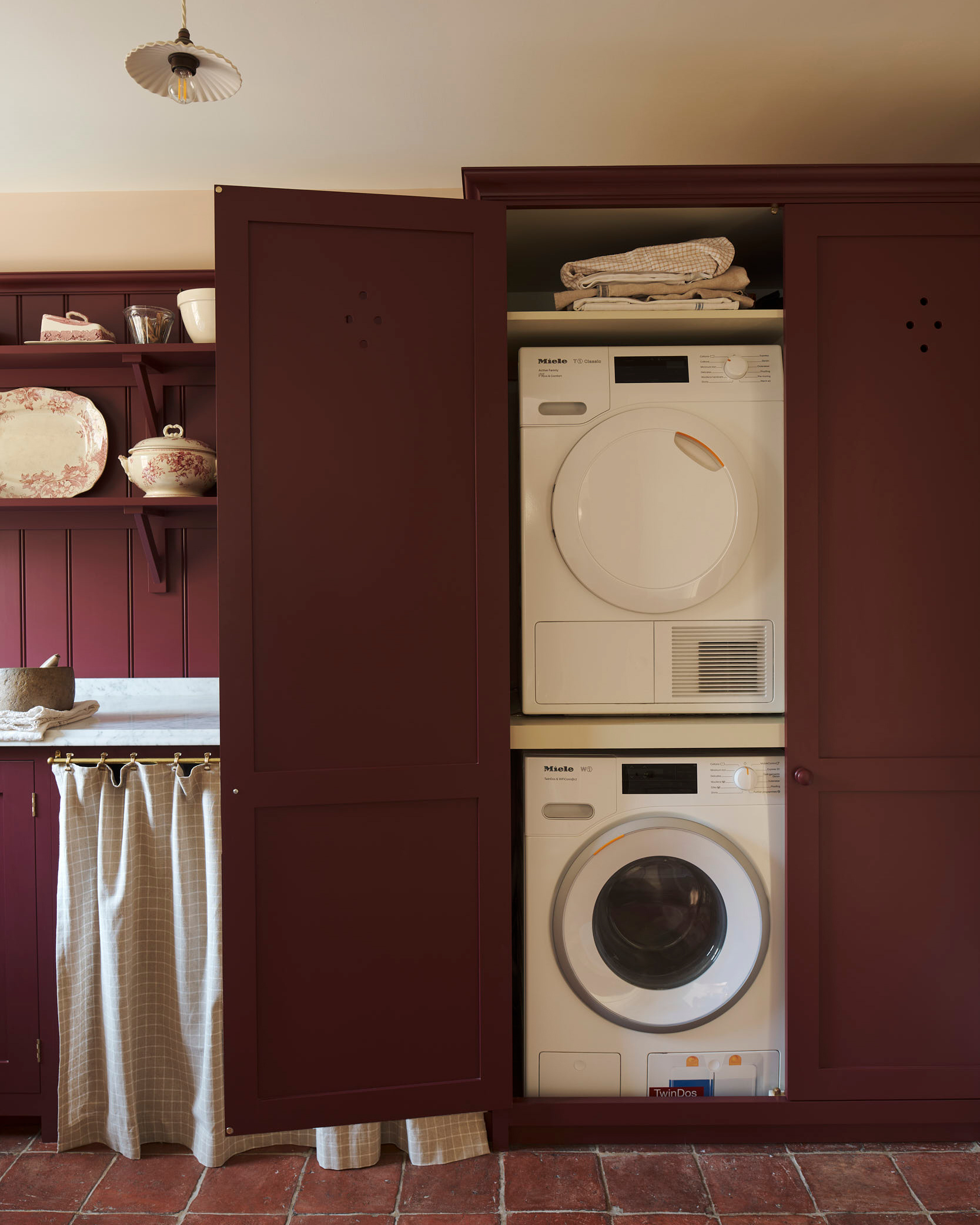 a utility room with stacked washing machines in a deep red cupboard, terracotta stone floors and open shelves