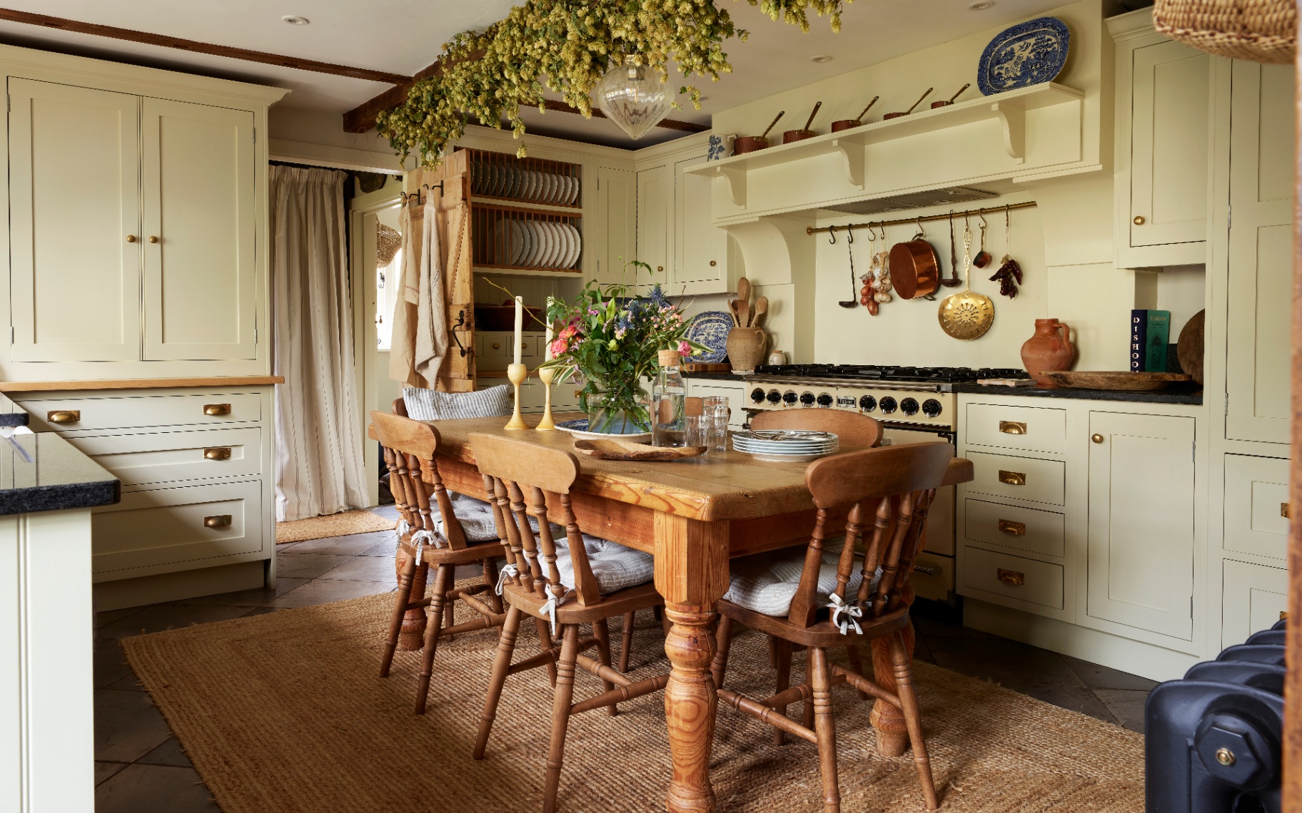 a cottage kitchen with pale green cabinetry, wooden beams, a large farmhouse table and chairs with a jute rug underneath