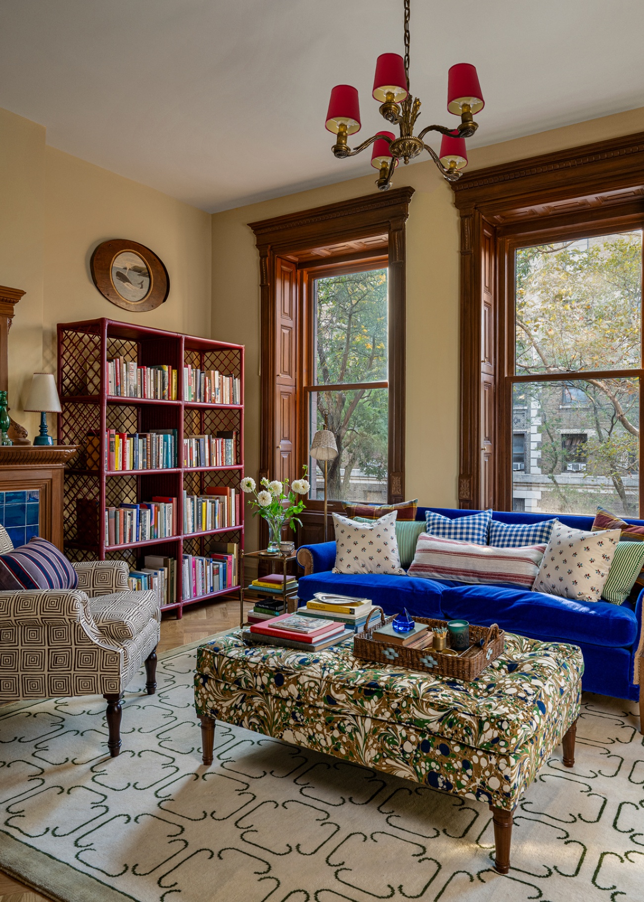 A library with a vibrant blue sofa, marbled upholstered ottoman, patterned armchair all arranged on a green-and-white rug, with a multi-arm chandelier above and a red lacquer bookshelf to the right. Two large windows with dark wood recesses are in the back