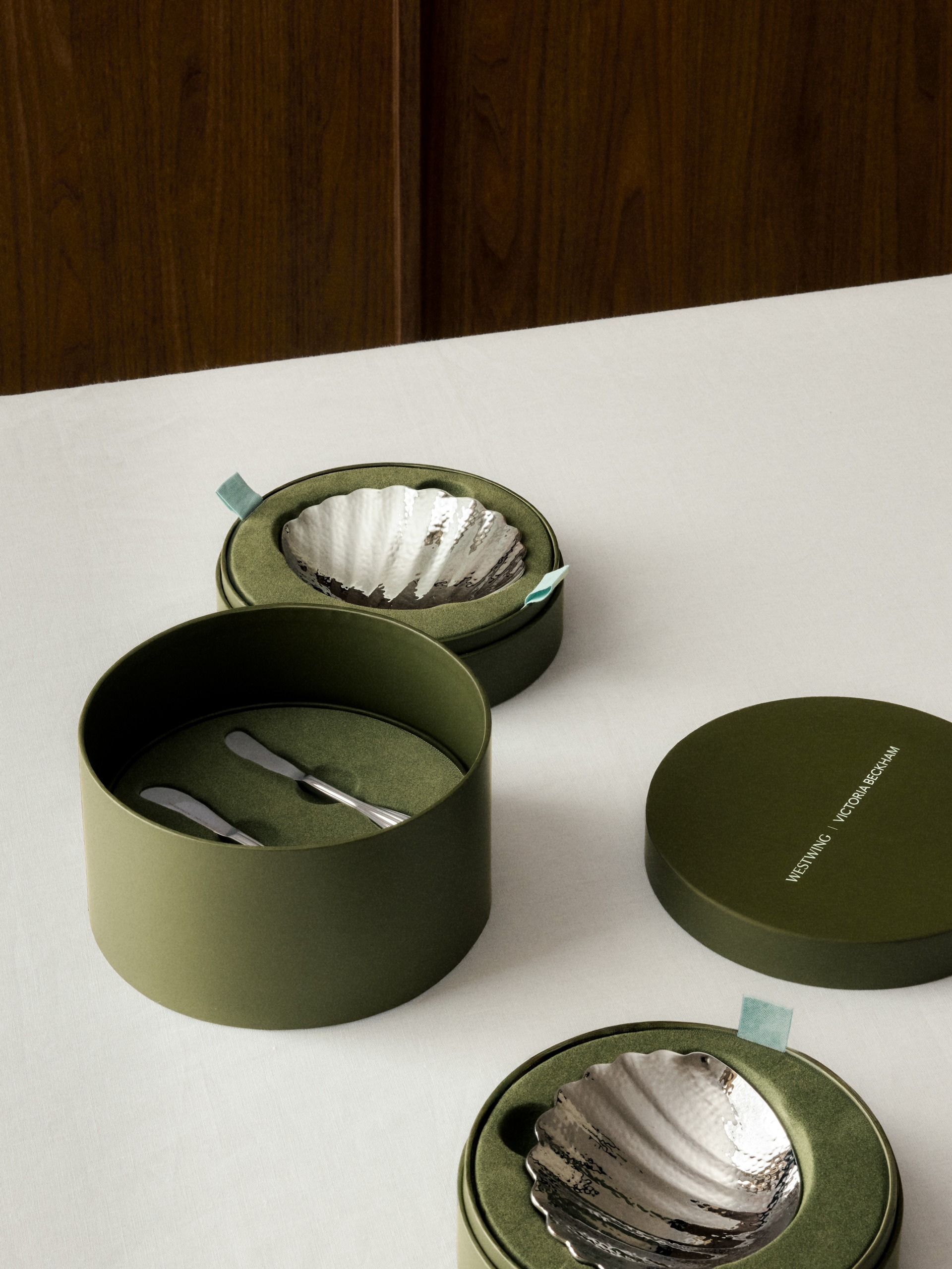 A photograph showing luxurious olive green gift packaging on a white tablecloth, displaying textured silver shell-shaped dishes and small butter knives nestled in velvet-lined boxes with natural wood behind