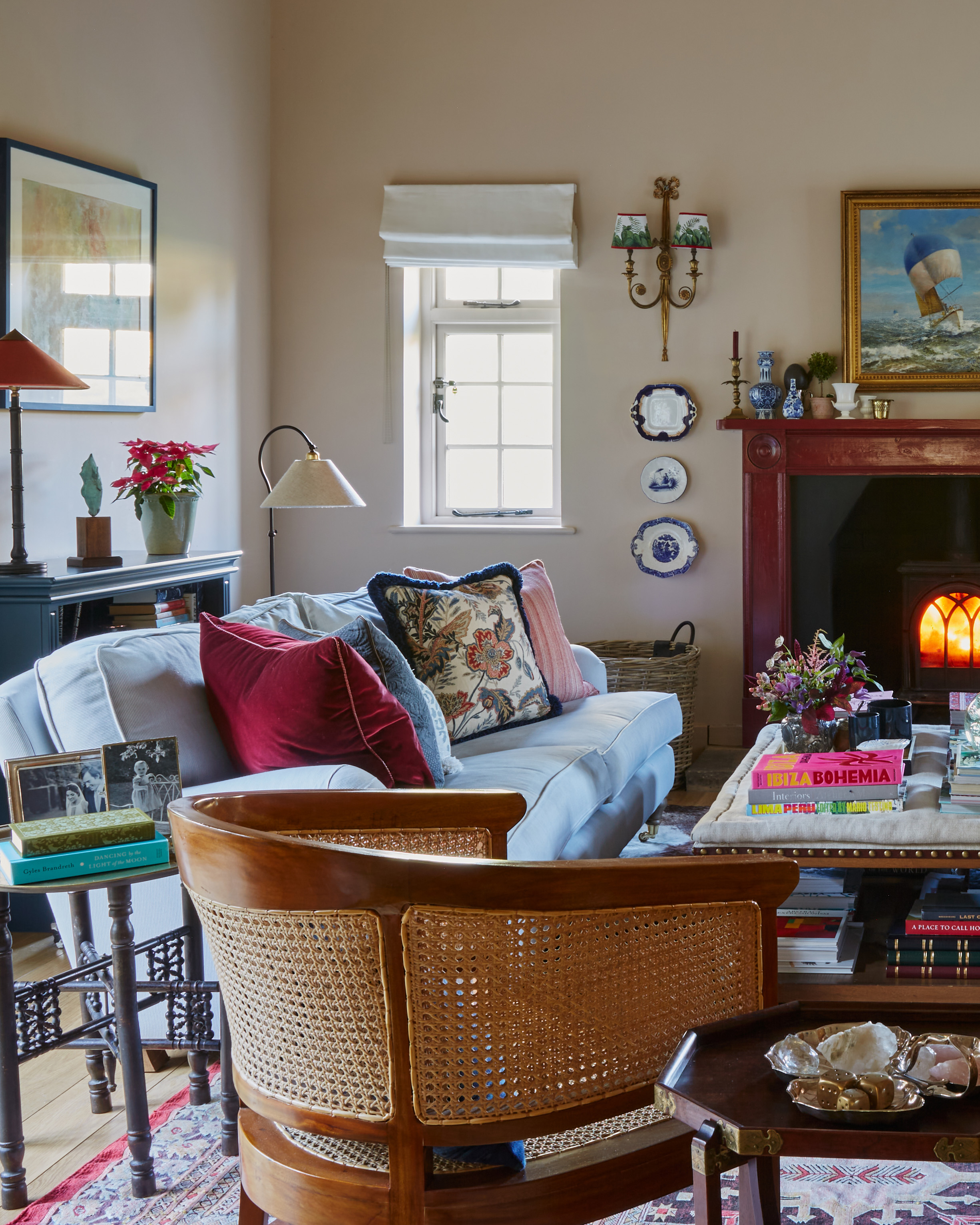 a living room with a lit wood burner in a fireplace, a cream ottoman coffee table filled with books and decor, a pale blue and white stripe sofa and a wooden and rattan arm chair