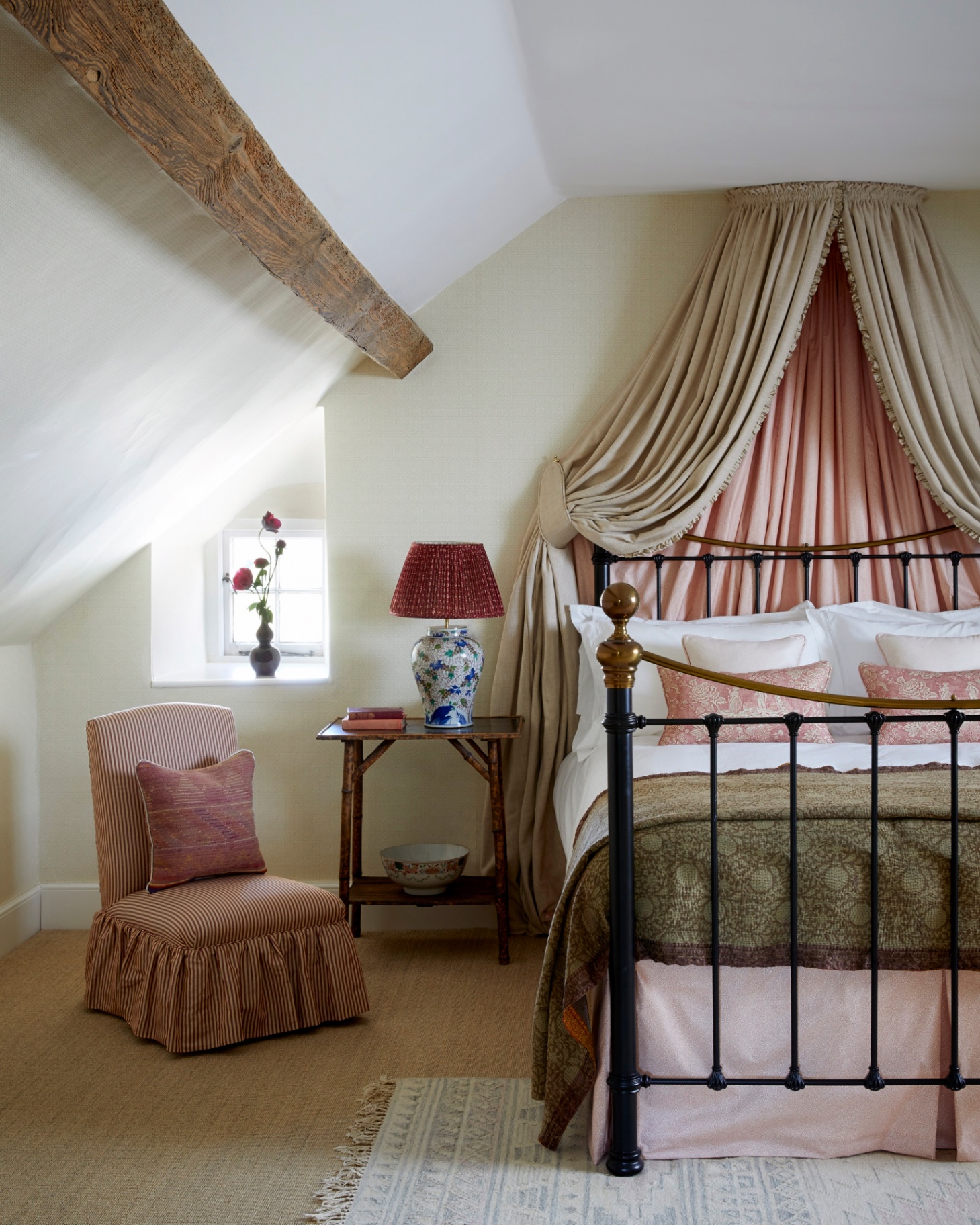 an eaves bedroom with wooden beams, a red adn white stripe skirted accent chair and a large bed with a cream and pink canopy, white bedding and an olive green bedspread
