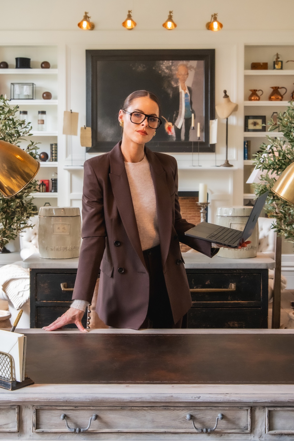 Jade Holland Cooper in a brown blazer holding a laptop in her home office, backed by styled bookshelves and brass lamps
